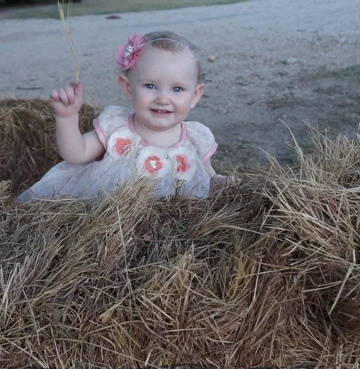 A young girl with blonde hair, blue eyes, and a pink headband with flowers, smiling and sitting behind a pile of hay at an outdoor setting.
