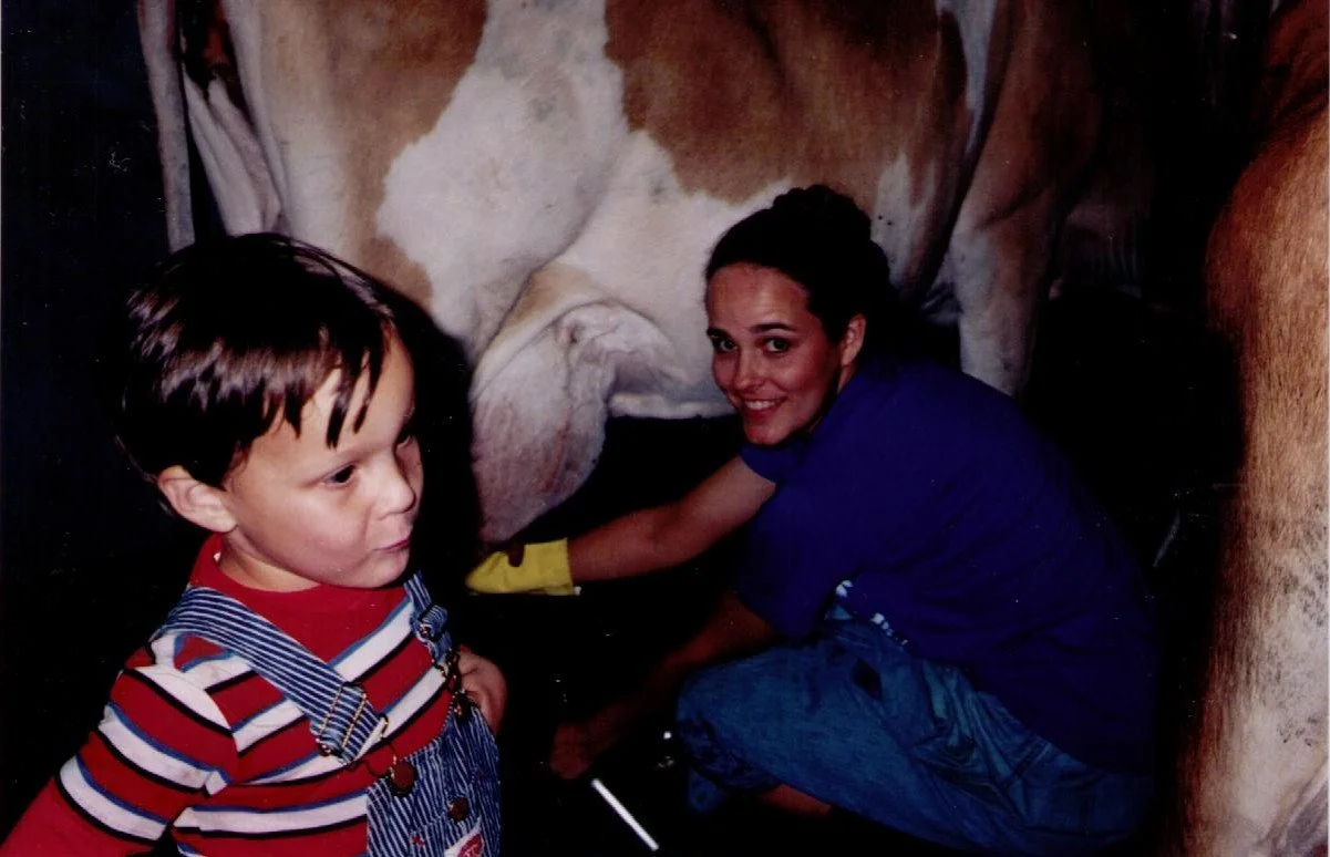 A woman and a young boy milking a cow inside a barn. The woman is smiling and wearing yellow gloves, while the boy is looking away.
