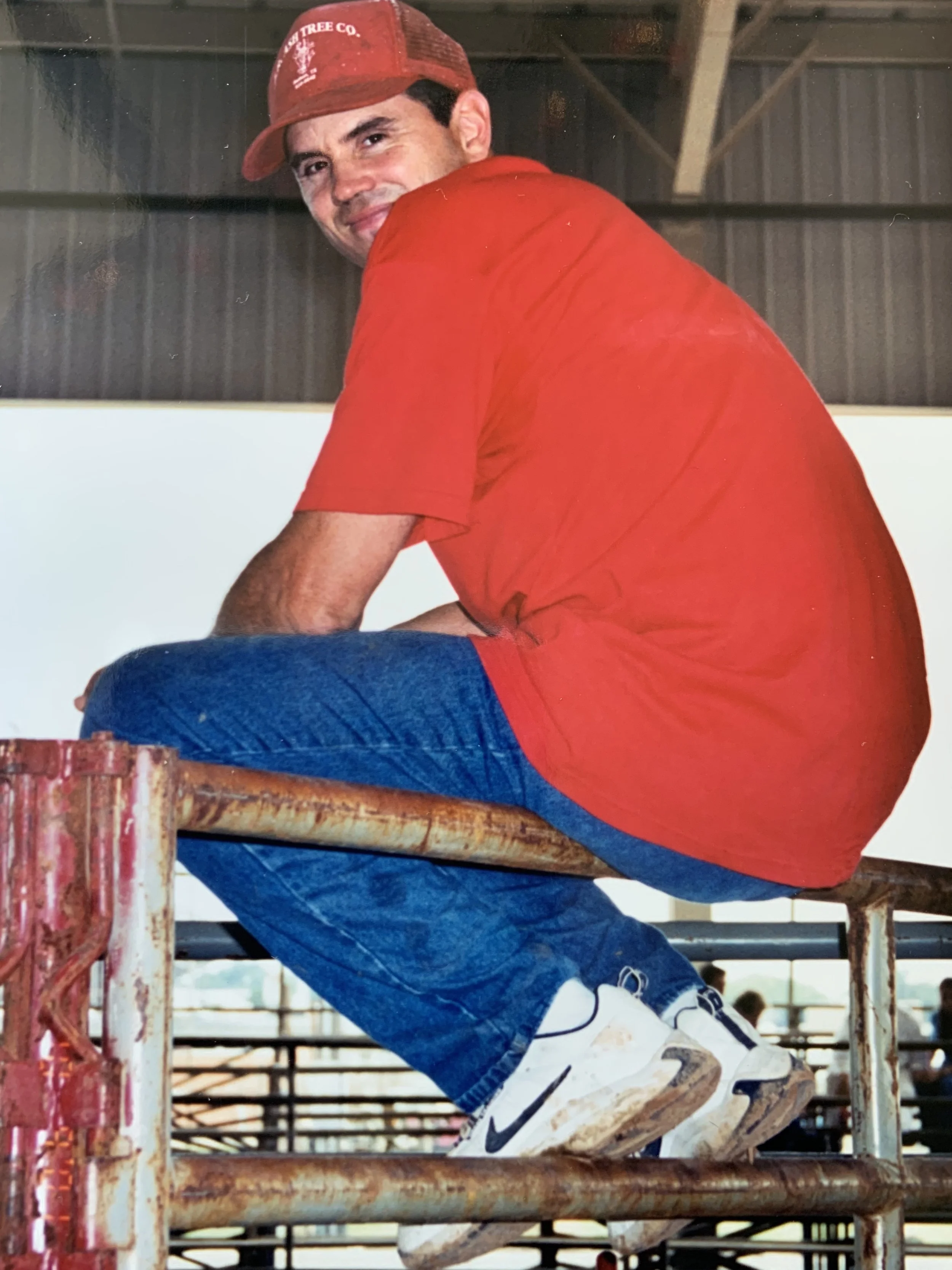 Man in red t-shirt and cap sitting on a rusted metal railing in a warehouse-like area.