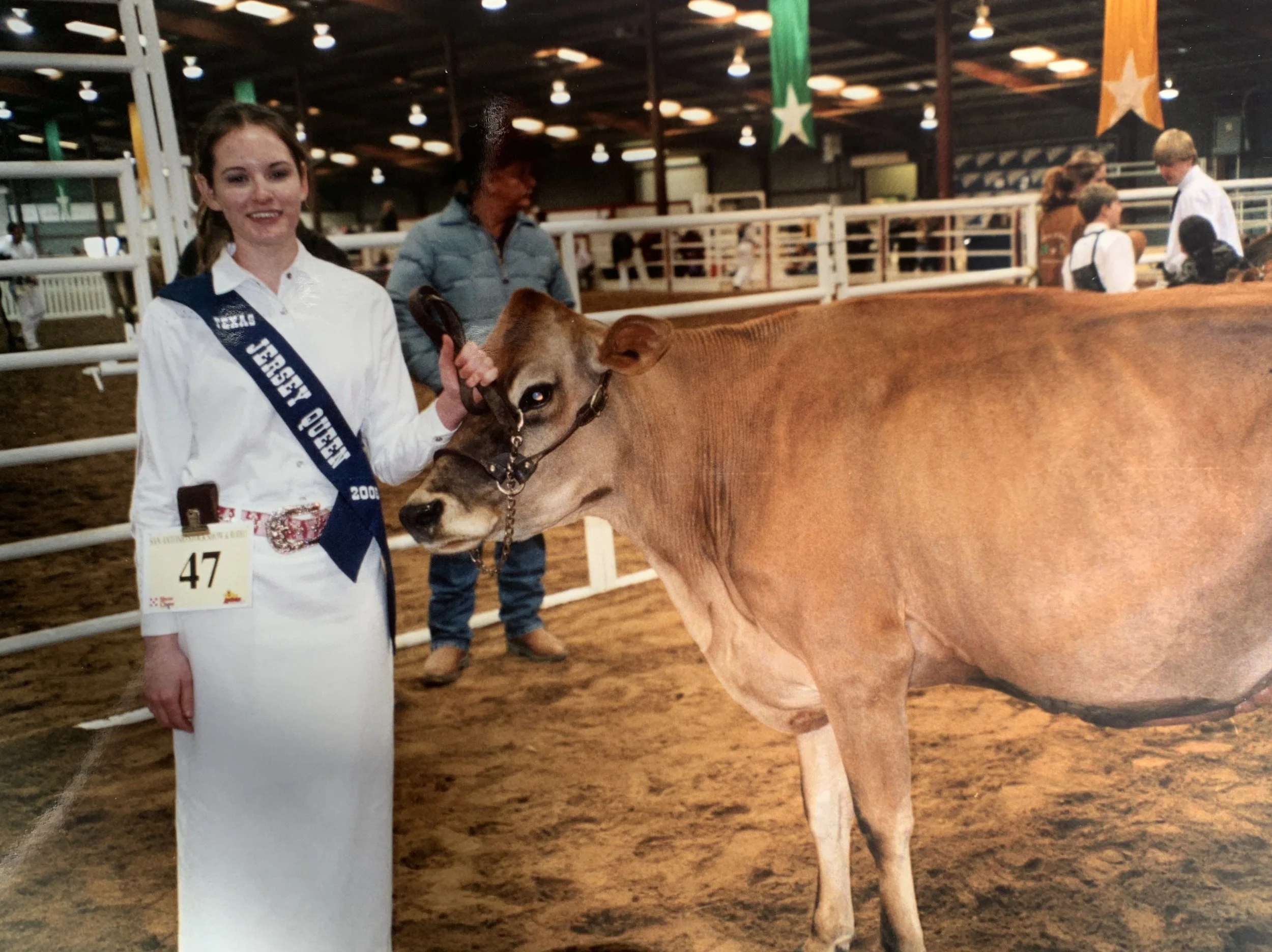 Young woman in a white dress with a sash and number 47 holding a cow's halter at a livestock exhibition inside a large barn with several people and colorful banners in the background.