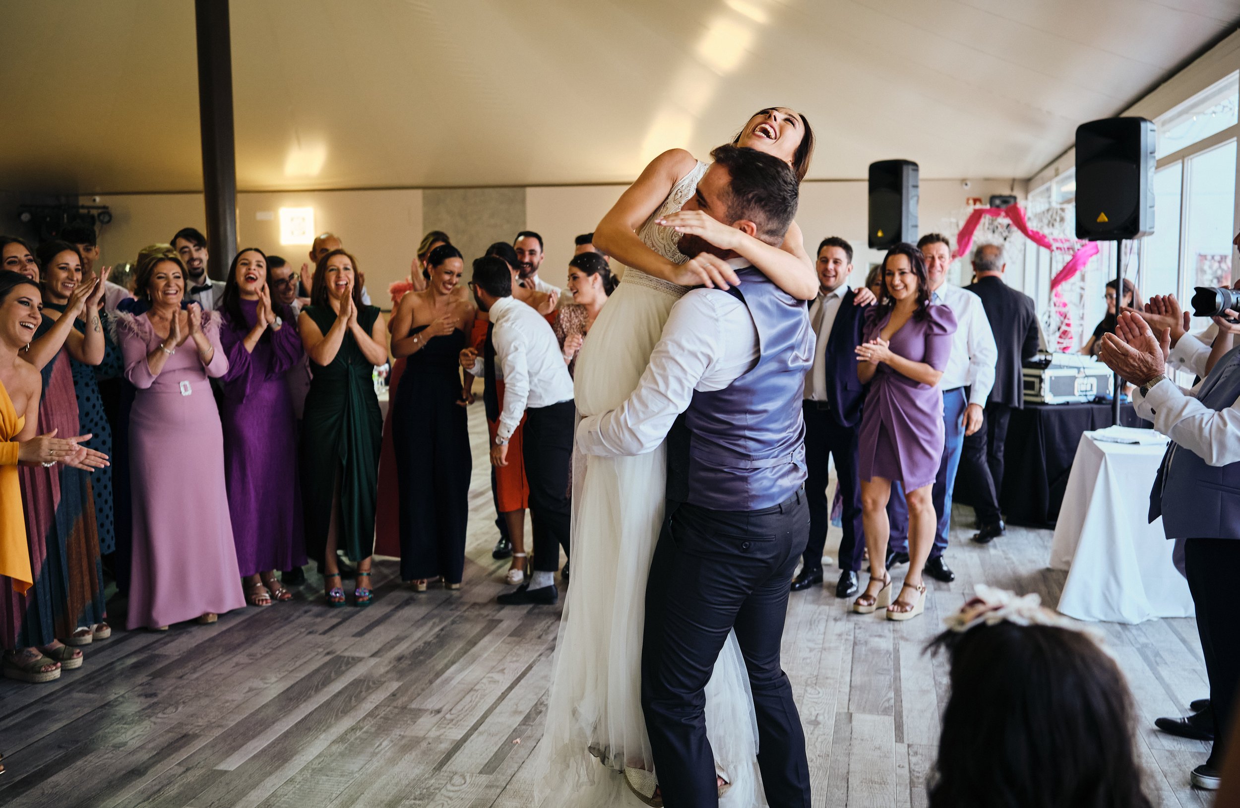 Celebración de boda con personas aplaudiendo, un novio sosteniendo a la novia mientras ella sonríe y invitados alrededor.