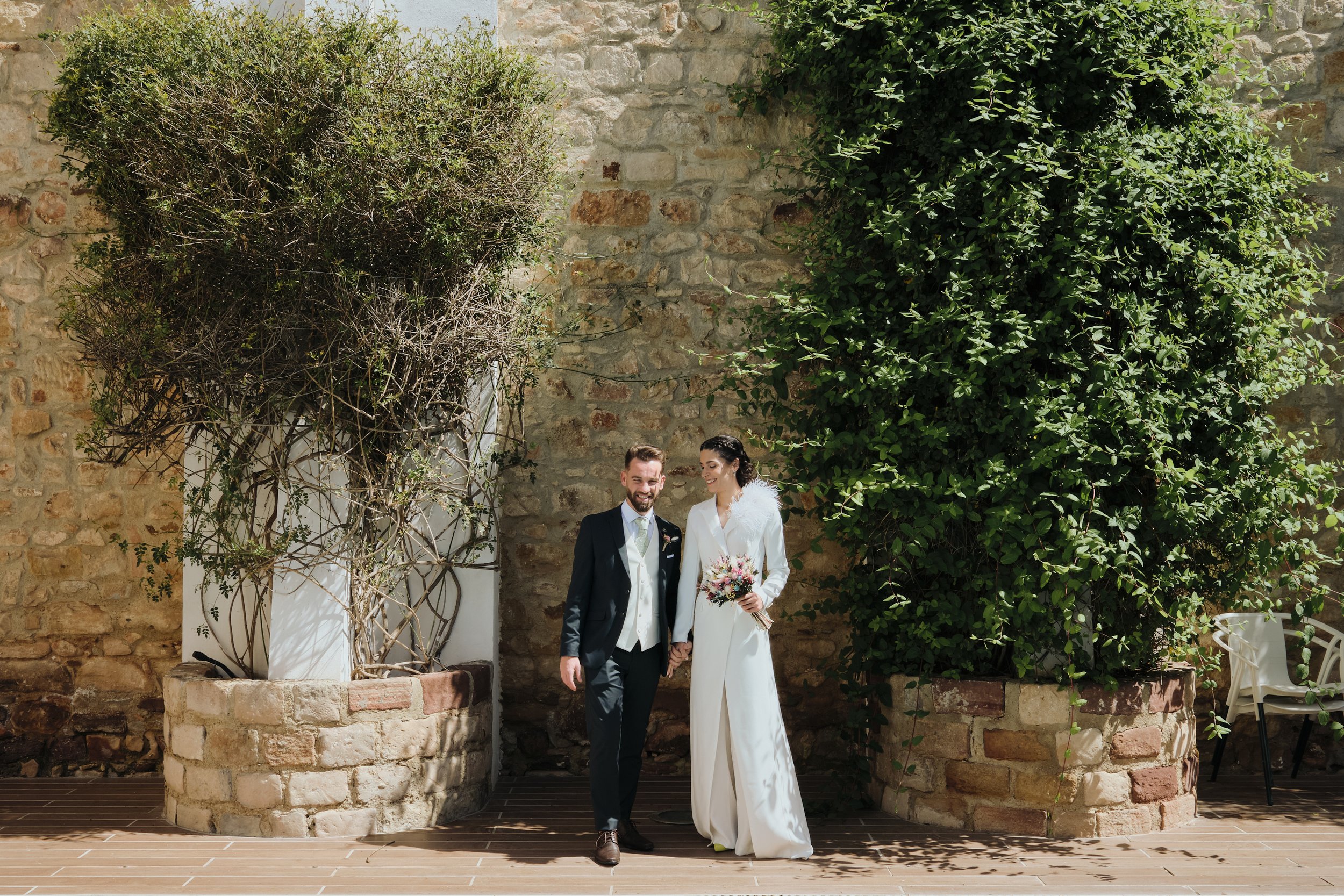 Pareja de recién casados caminando en un jardín con muros de piedra y plantas.