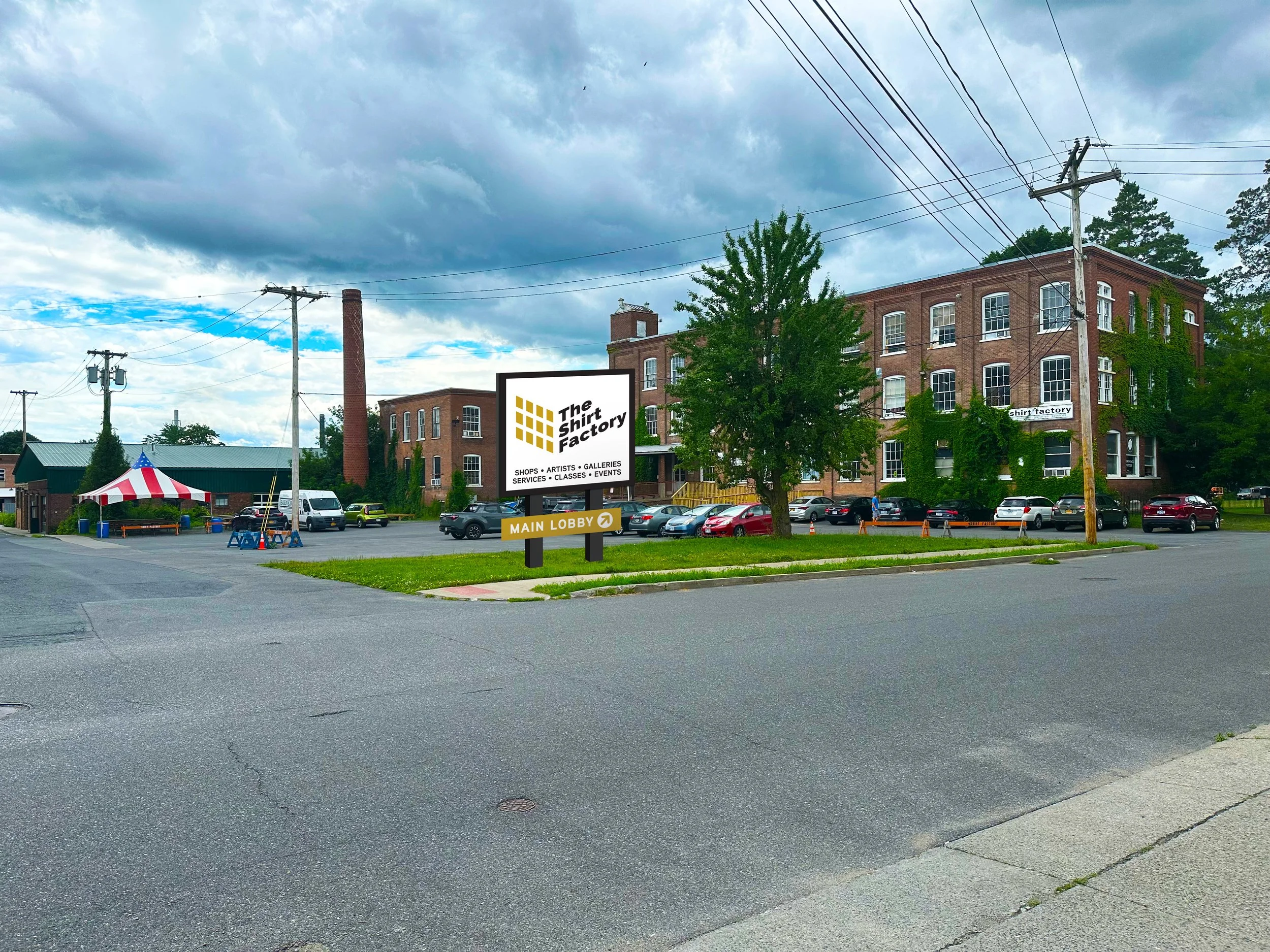 Brown brick building labeled 'The Shirt Factory' with parking lot and cars in the foreground. A red and white tent structure is visible to the left.