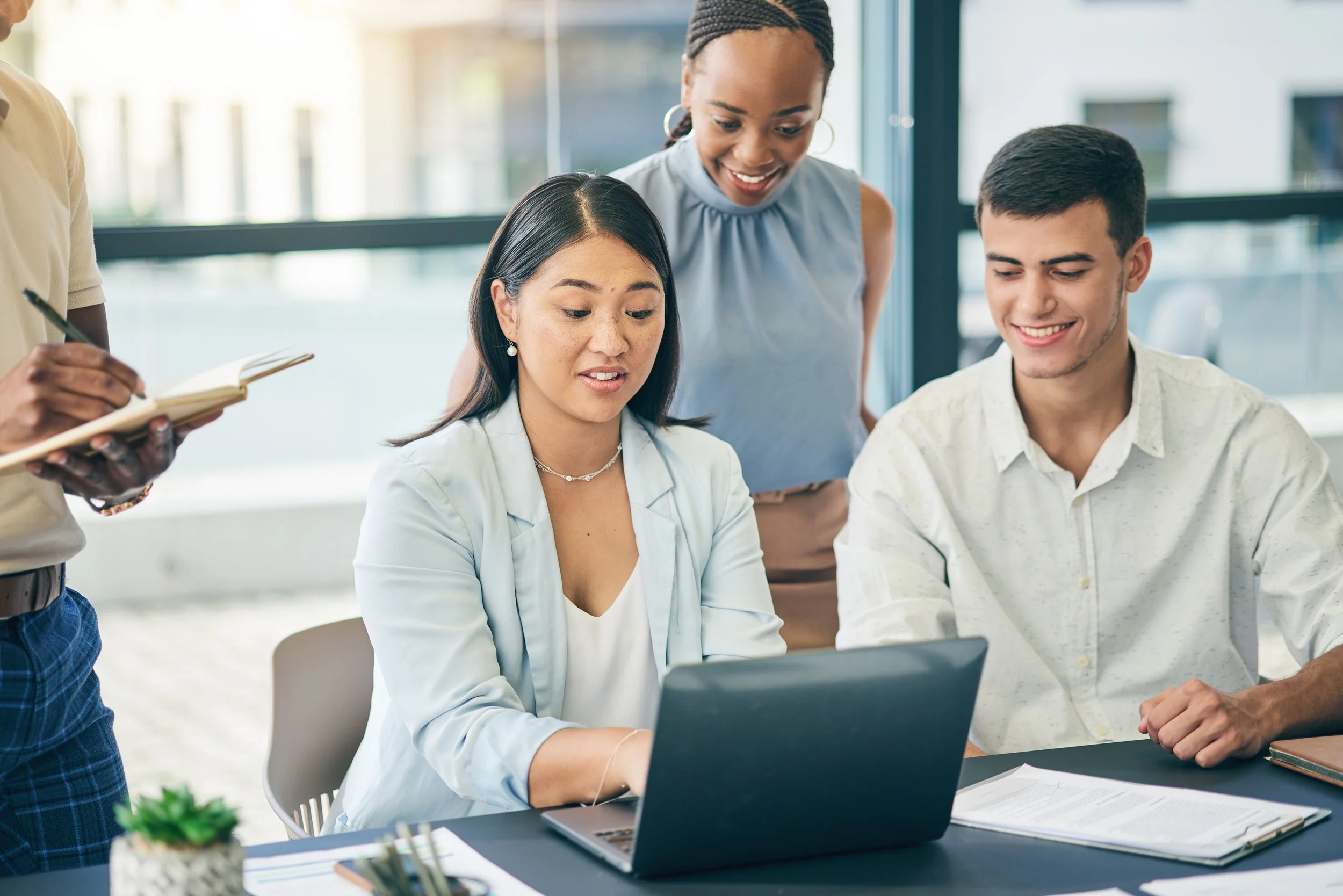 A diverse group of five professionals in a modern conference room, engaged in a discussion. One person is standing and smiling, holding a paper, while others are seated with laptops and notes, some smiling and listening attentively.