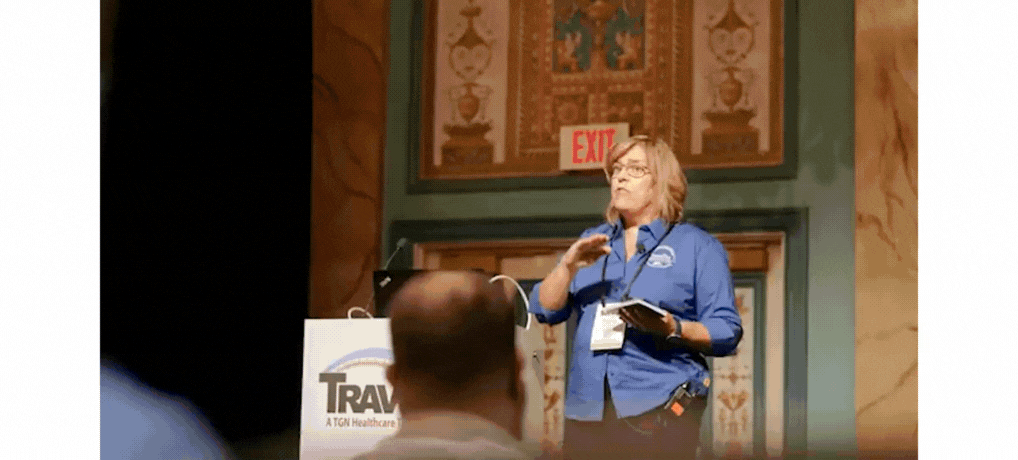 A woman with glasses, wearing a blue shirt, giving a presentation at a conference with a TGN Healthcare banner, an attendee's head visible in the foreground, and an exit sign behind her.