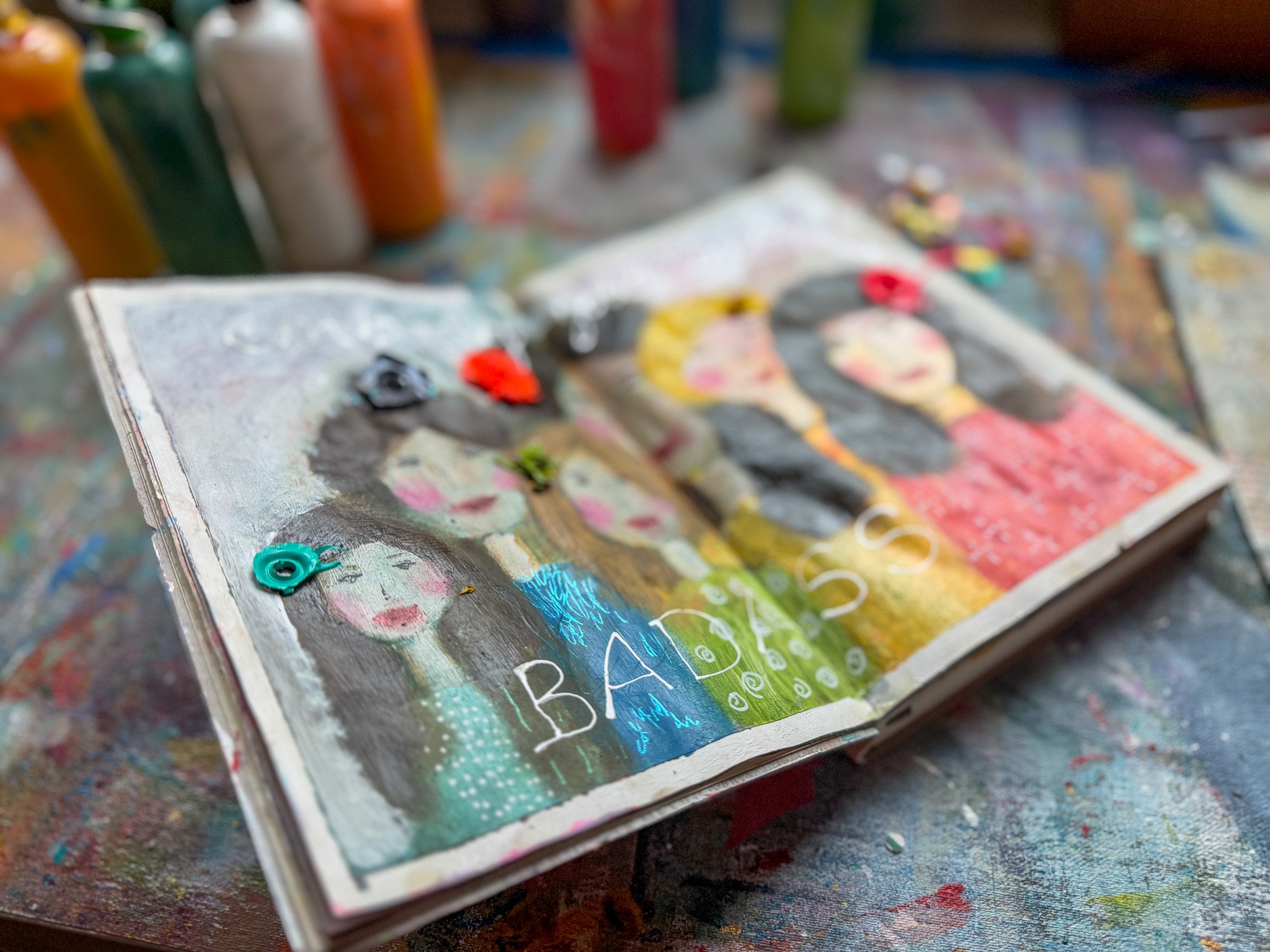 An open art journal features a painted illustration of five women with dark hair, with the word "BADASS" at the bottom, surrounded by paint bottles and art supplies on a colorful, paint-splattered table.