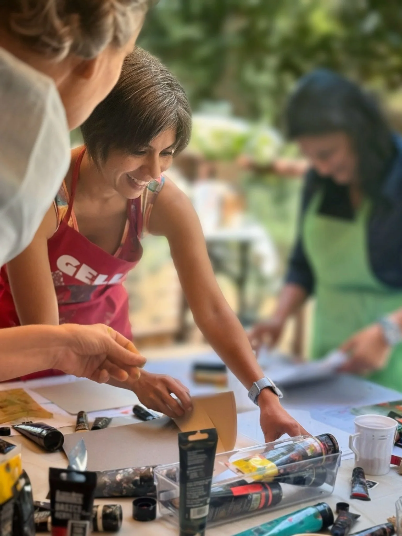 People making art or craft projects at a table outdoors, with various art supplies like paint tubes, brushes, and containers scattered on the table.