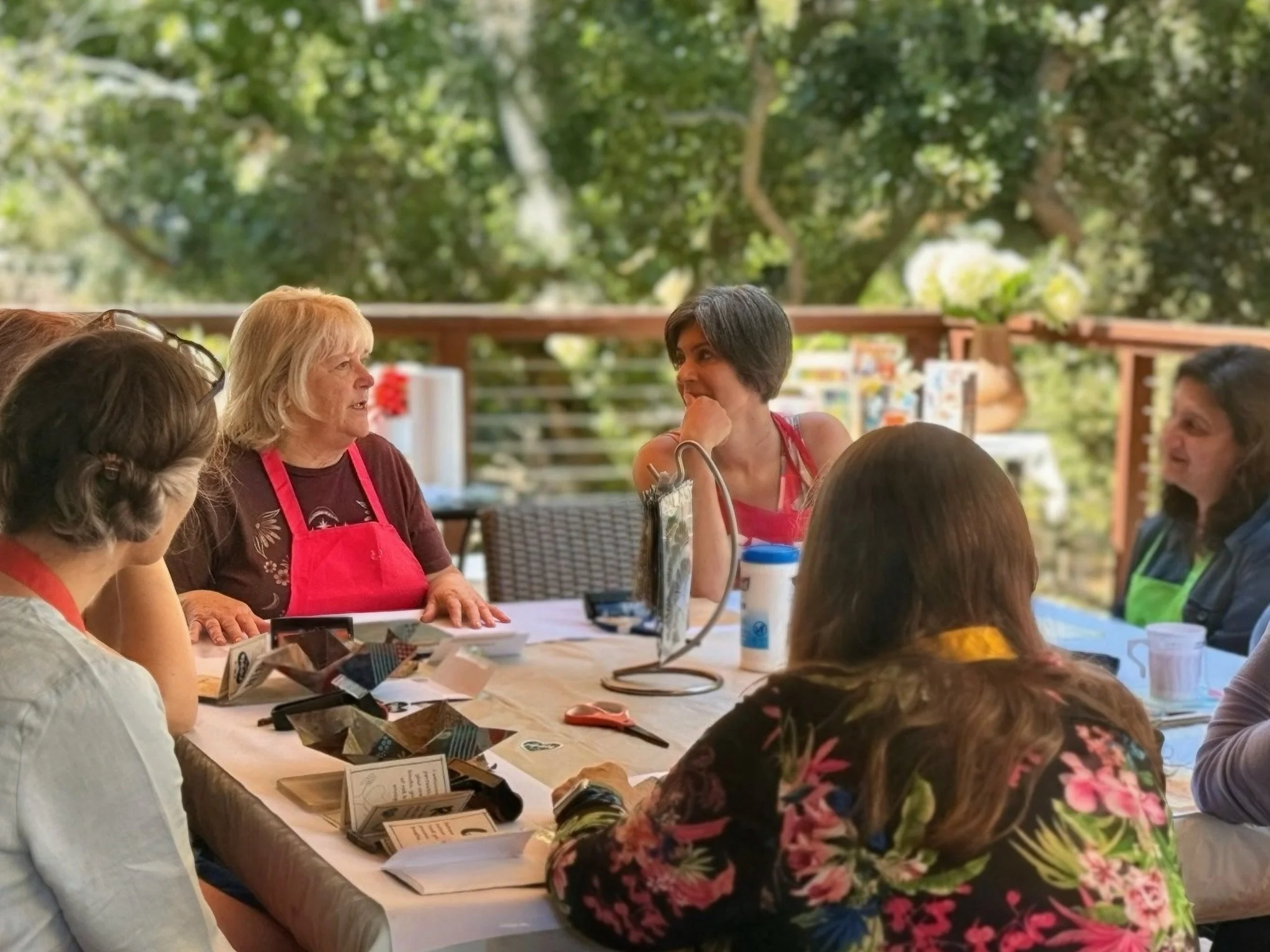 Group of women sitting around a table outdoors, engaging in conversation during a garden gathering or workshop.