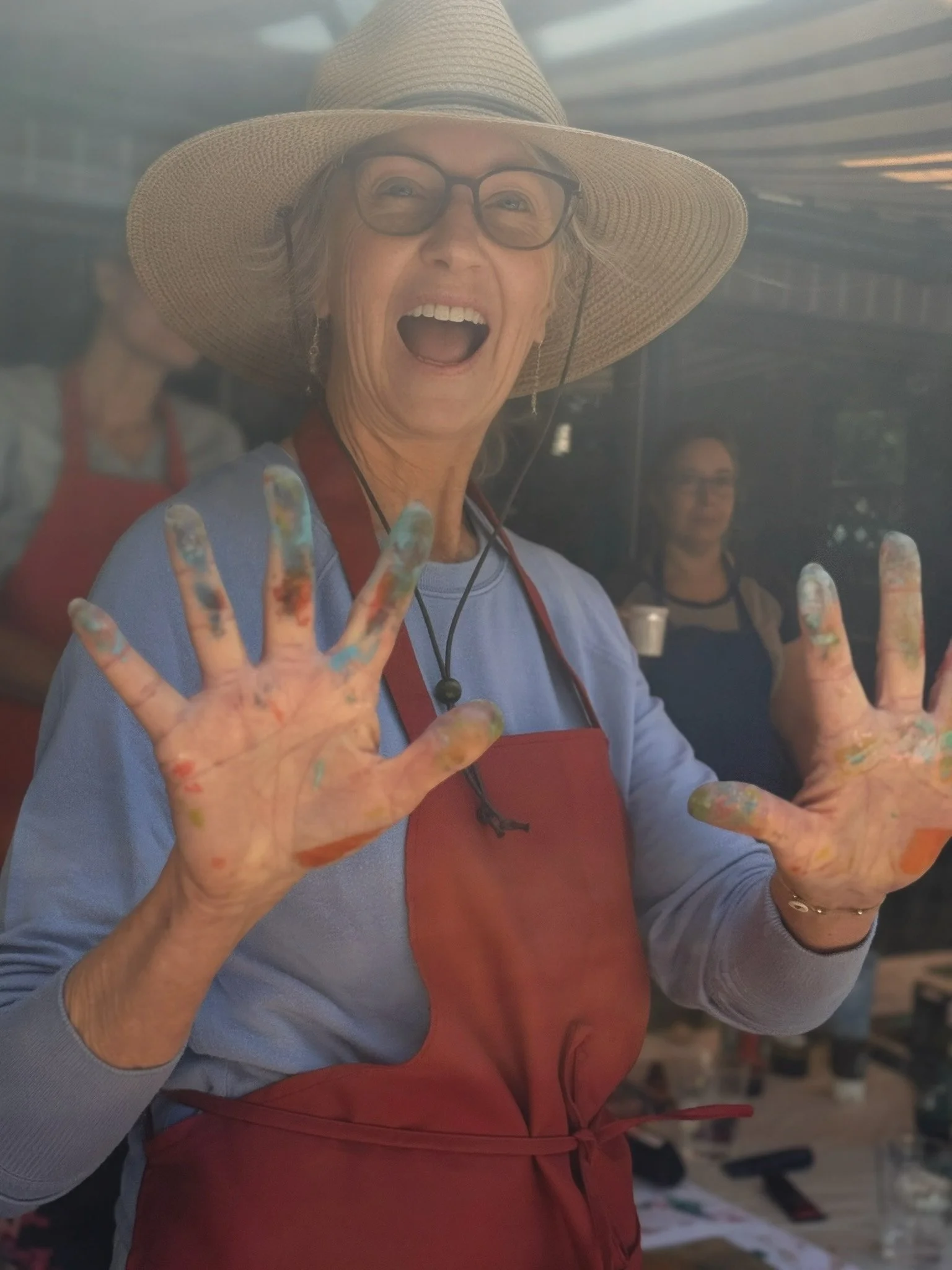 A woman wearing a large straw hat, glasses, and a red apron, smiling with her mouth open, showing her painted fingers raised in a peace sign gesture.