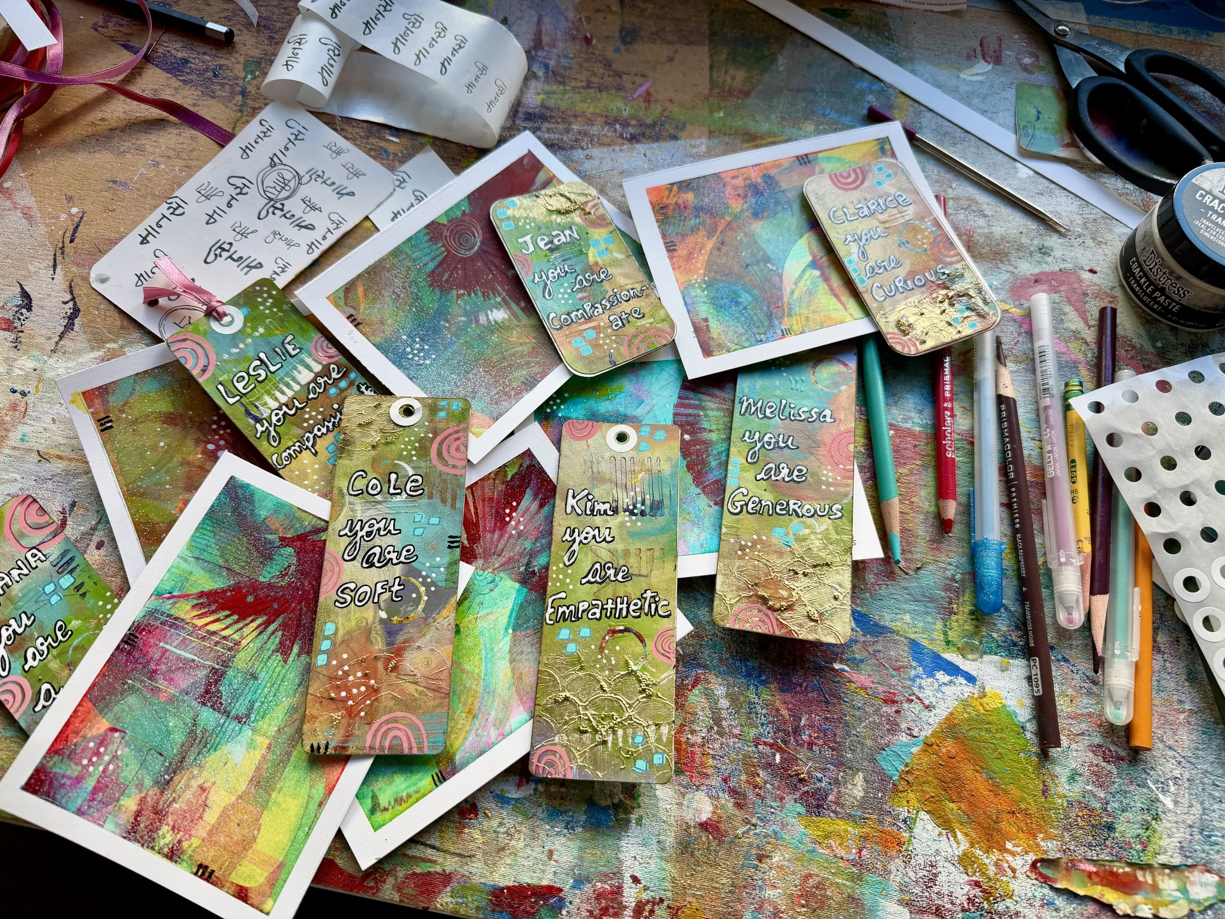 A colorful, abstract art workspace with handmade tags, papers, and art supplies like pens, scissors, and paint on a splattered, multicolored table.