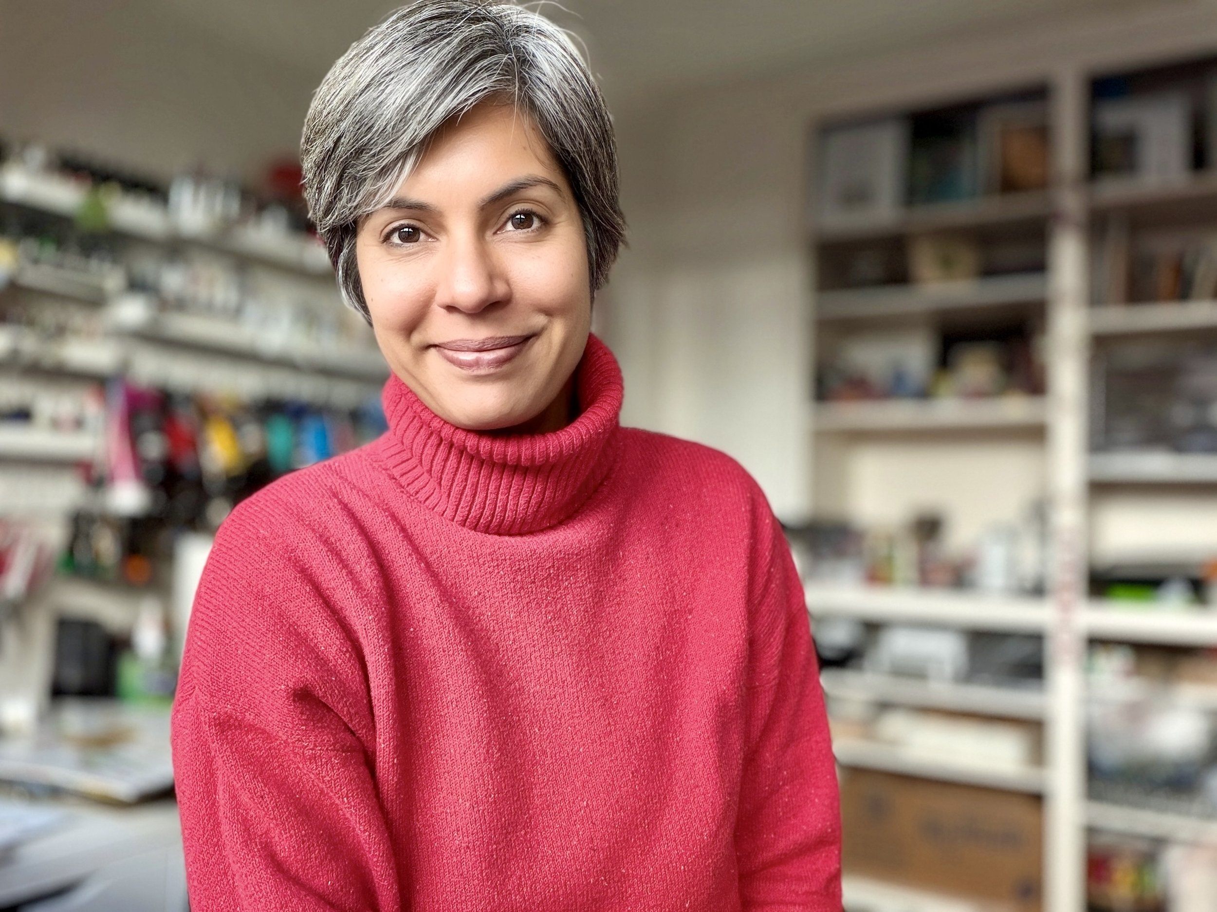 A woman with short gray hair wearing a red turtleneck sweater smiling in an indoor space with shelves filled with books and miscellaneous items in the background.
