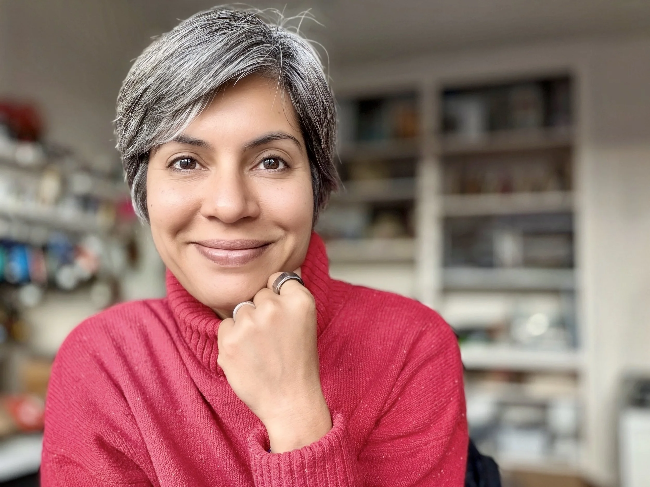 A smiling woman with short gray hair wearing a red sweater, seated in a room with blurred shelves in the background.