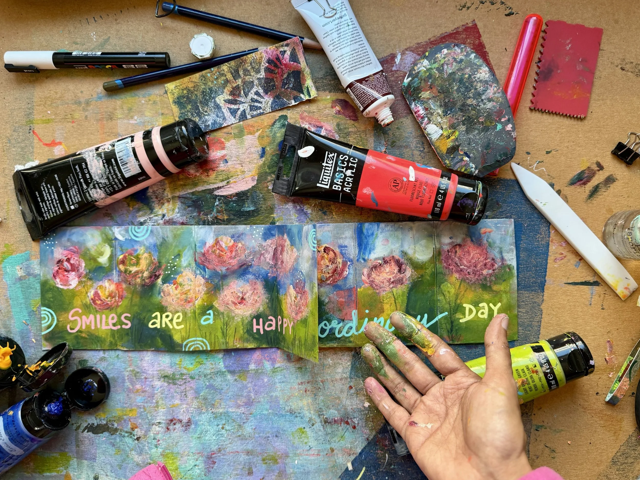 Colorful art project on paper with handwritten words "Smiles are a happy ordering day" surrounded by paint supplies, including tubes of paint, brushes, a palette, and paint on the workspace.