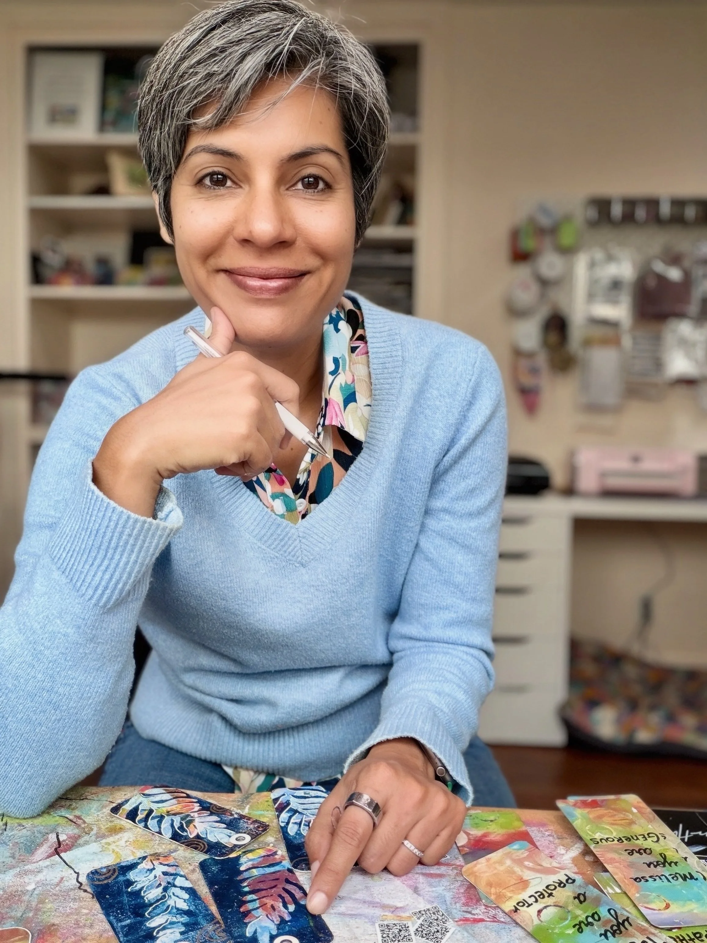 A woman with short gray hair wearing a blue sweater sitting at a table with colorful cards, pointing at one of the cards.