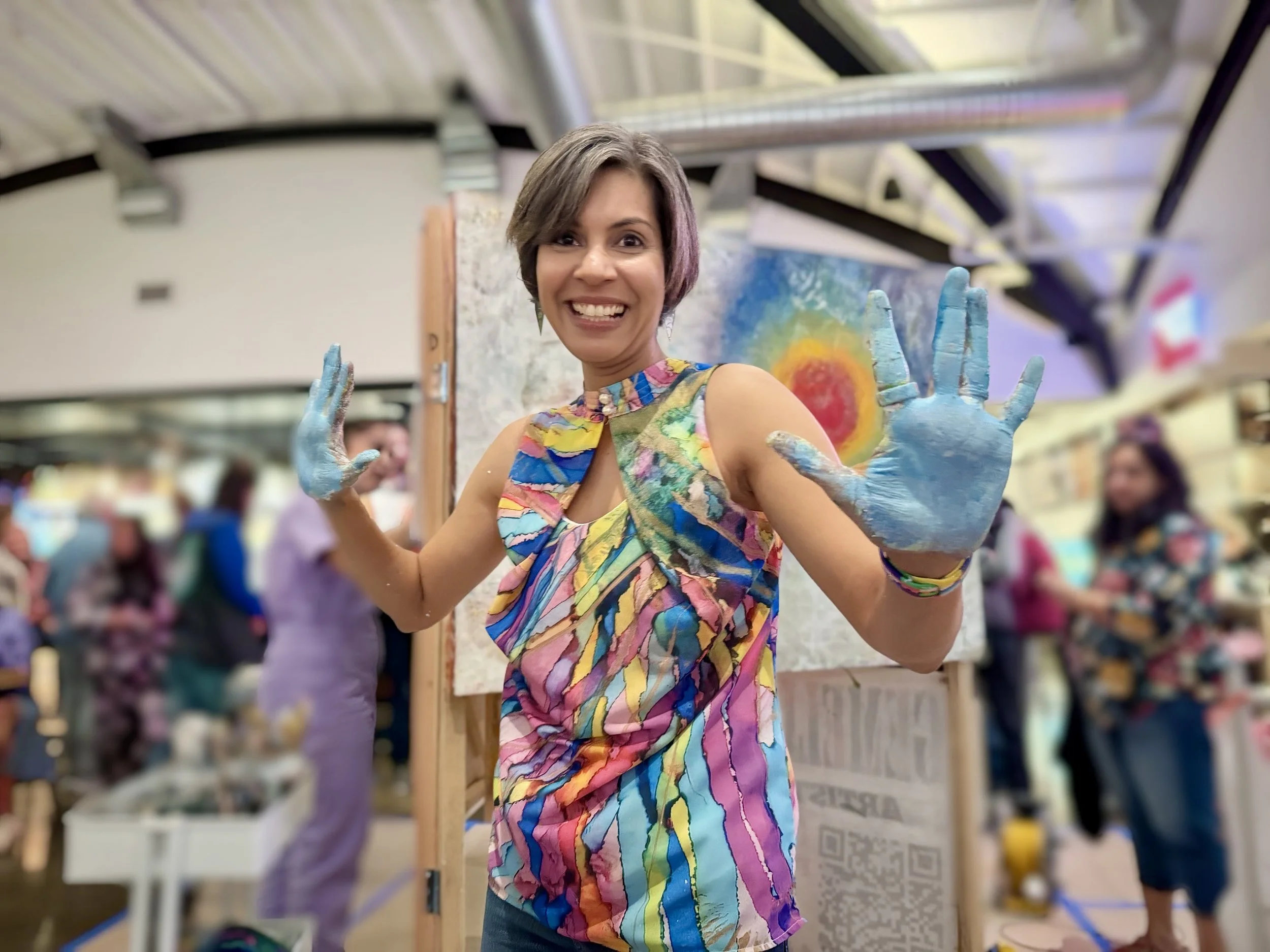 Mansi smiling with painted hands at an indoor art event, Live Painting Art Battle at State Street Market in Los Altos, colorful abstract artwork behind her, blurred crowd in background.