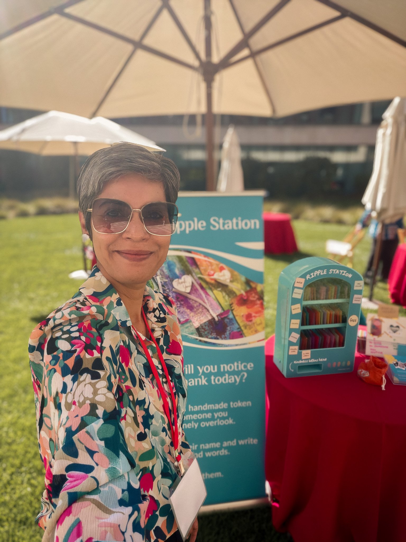 A woman with short gray hair wearing sunglasses and a colorful floral shirt stands outdoors at a booth with a sign that reads 'Ripple Station' during daytime. The booth appears to be for a community or charity event, with umbrellas, tables, and informational materials visible.