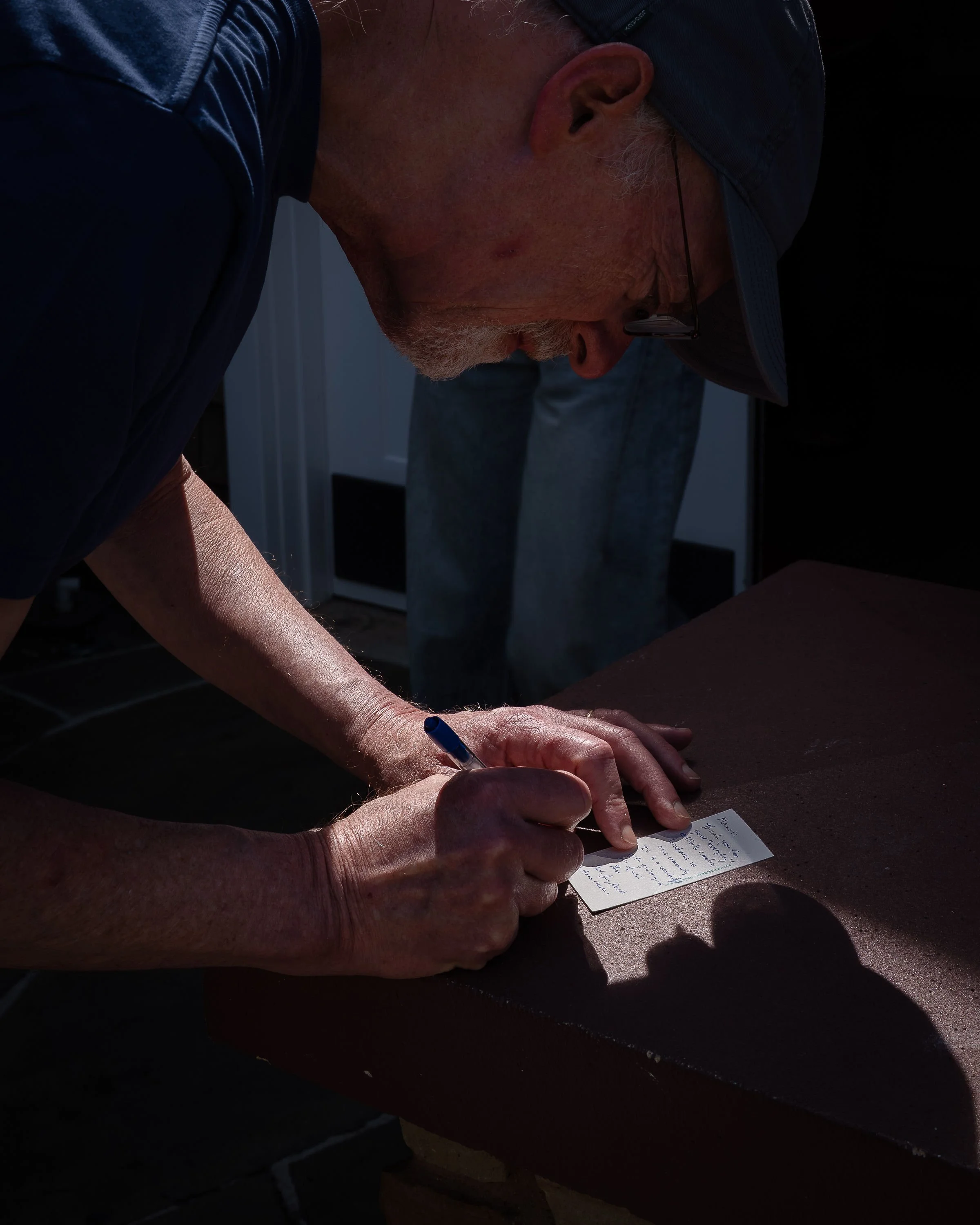 A man wearing a dark baseball cap and glasses is writing on a small piece of paper placed on a table. The focus is on his hands and the paper, with his face in profile and slightly blurred in the background.