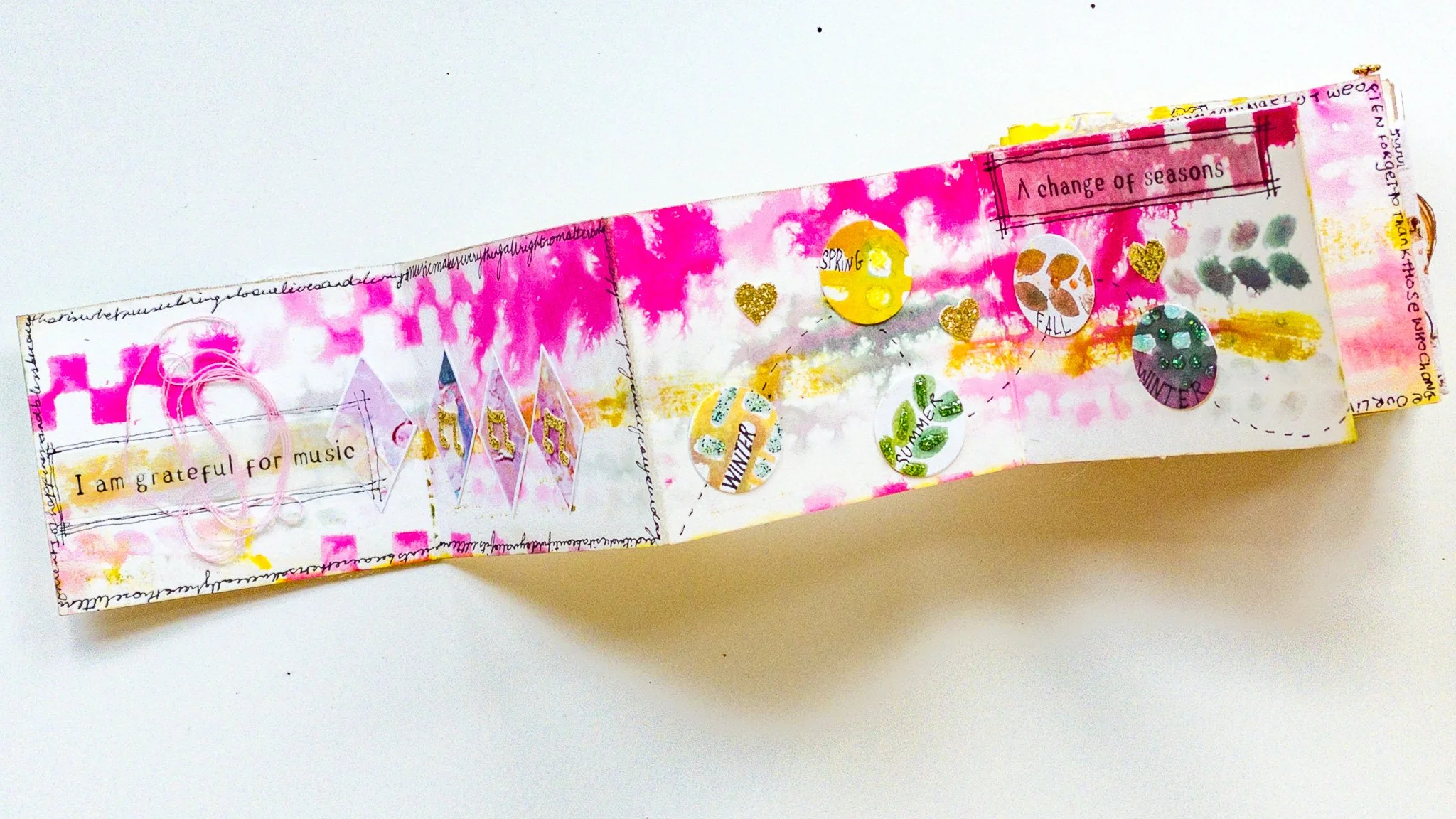 A colorful handmade scrapbook page with pink, yellow, and orange watercolor background, decorated with stickers, doodles, and the phrases 'I am grateful for music,' 'A change of seasons,' and seasonal words like 'Summer,' 'Fall,' 'Winter,' and 'Spring'.