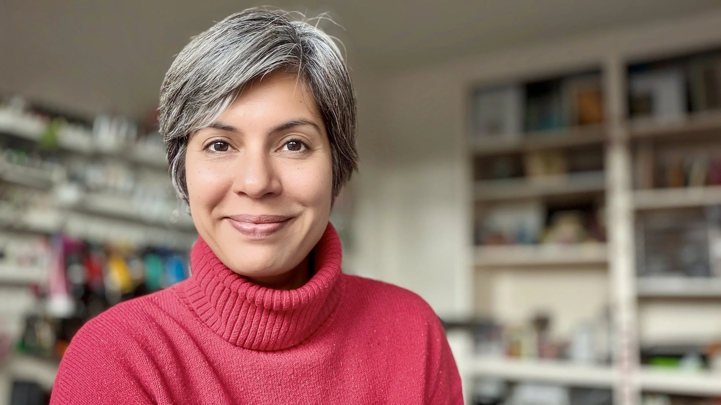 A smiling woman with short gray hair wearing a red turtleneck sweater, standing in a room with shelves filled with books and various items in the background.