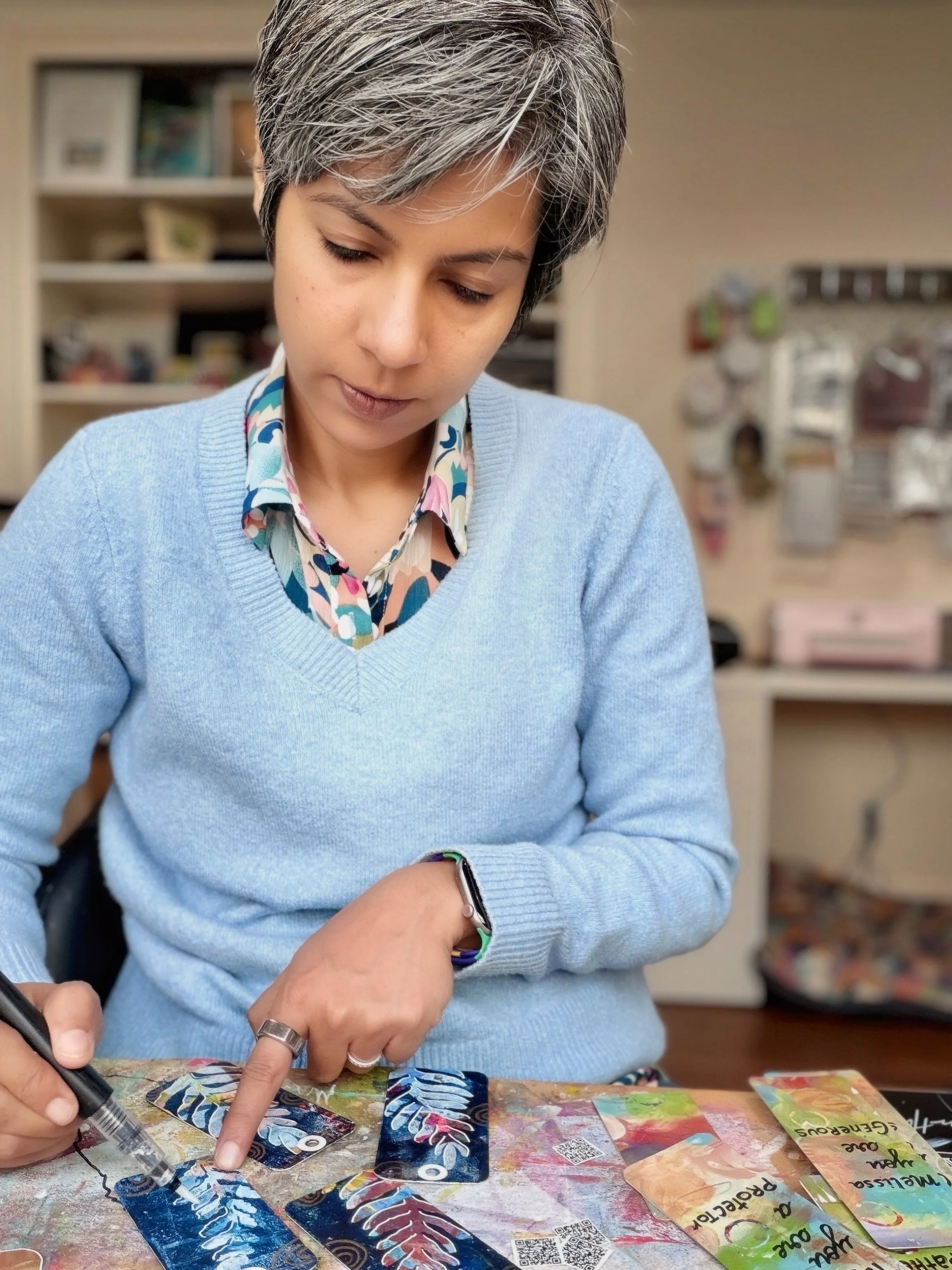 A woman with short, gray hair wearing a light blue sweater and a colorful patterned shirt is sitting at a table, examining and pointing to several cards with colorful, abstract designs, while holding a black marker.