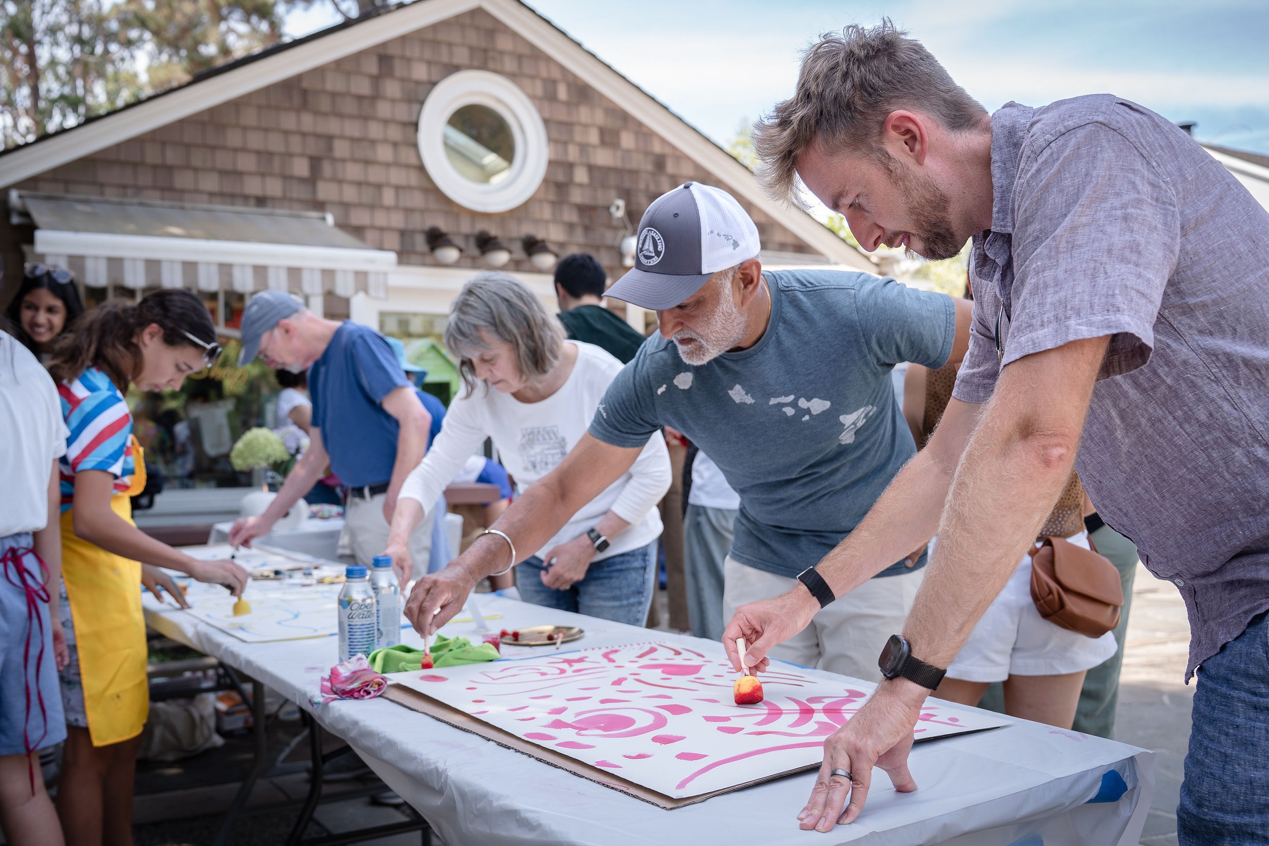Group of people participating in a community art project outdoors, painting on a large canvas laid on a table, with a house and trees in the background.