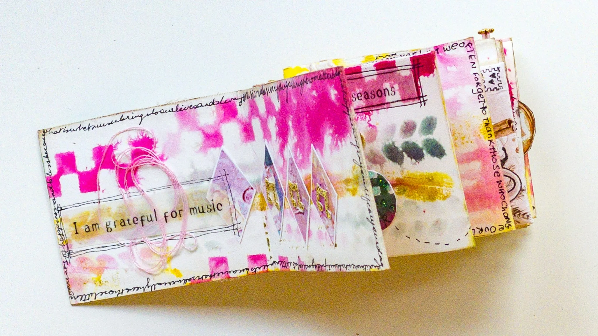 Close-up of a mixed media art journal spread with pink, yellow, and black watercolor splashes, handwritten text, and collaged elements, including the phrase "I am grateful for music."