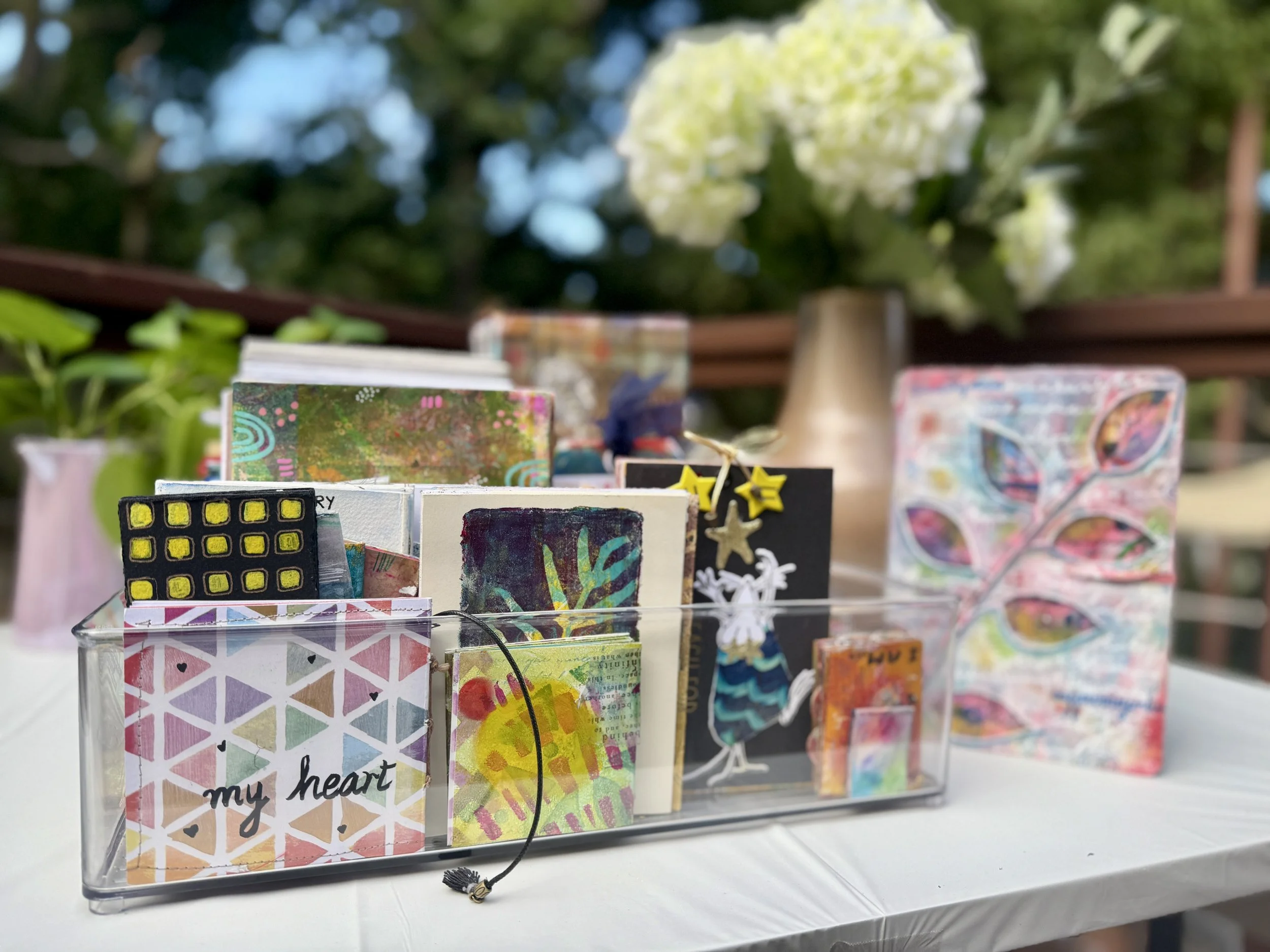 Assorted handmade cards with colorful, artistic designs inside a clear acrylic box on a white table, with a large white flower in a vase and blurred greenery in the background.