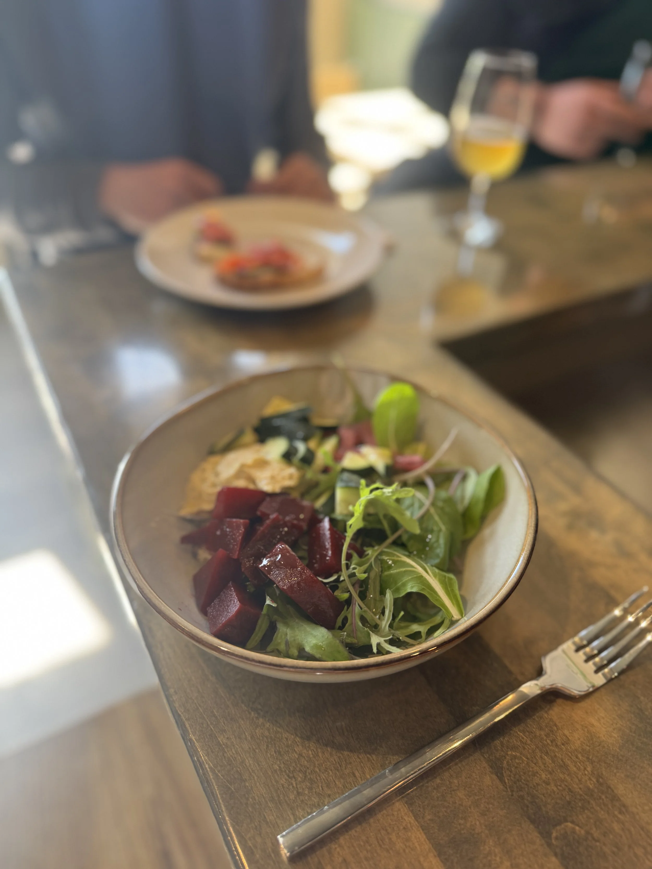 people sitting at a bar with food. There is a bowl with grains, greens, hummus and beets. Smoked salmon tartine in the background