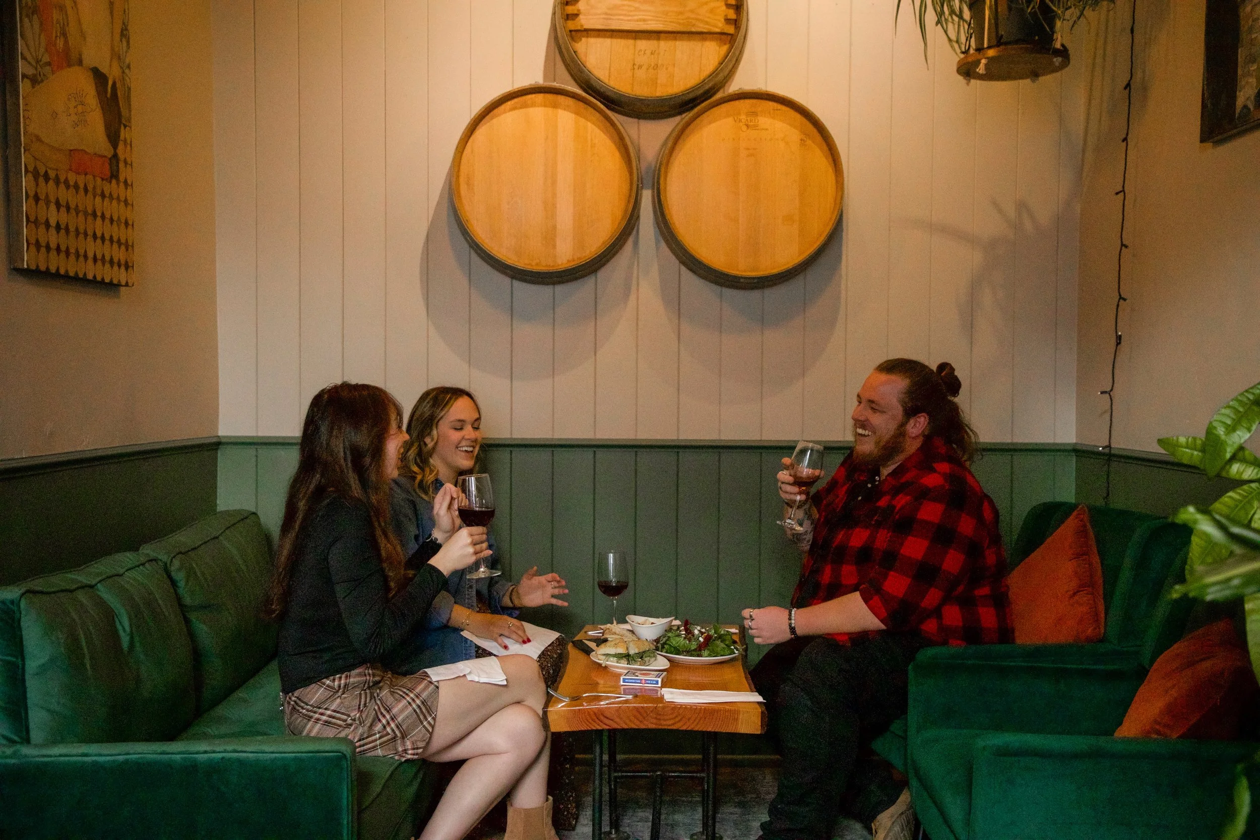 Four friends are sitting on a green booth in a restaurant, laughing and sharing drinks. The table has salads and wine glasses. The walls are white with three round wooden wine barrel decorations and some artwork. One woman is wearing a black top and plaid skirt, the woman next to her is in a dark jacket, and the man is wearing a red and black flannel shirt with a bun hairstyle. They appear to be enjoying a lively conversation.