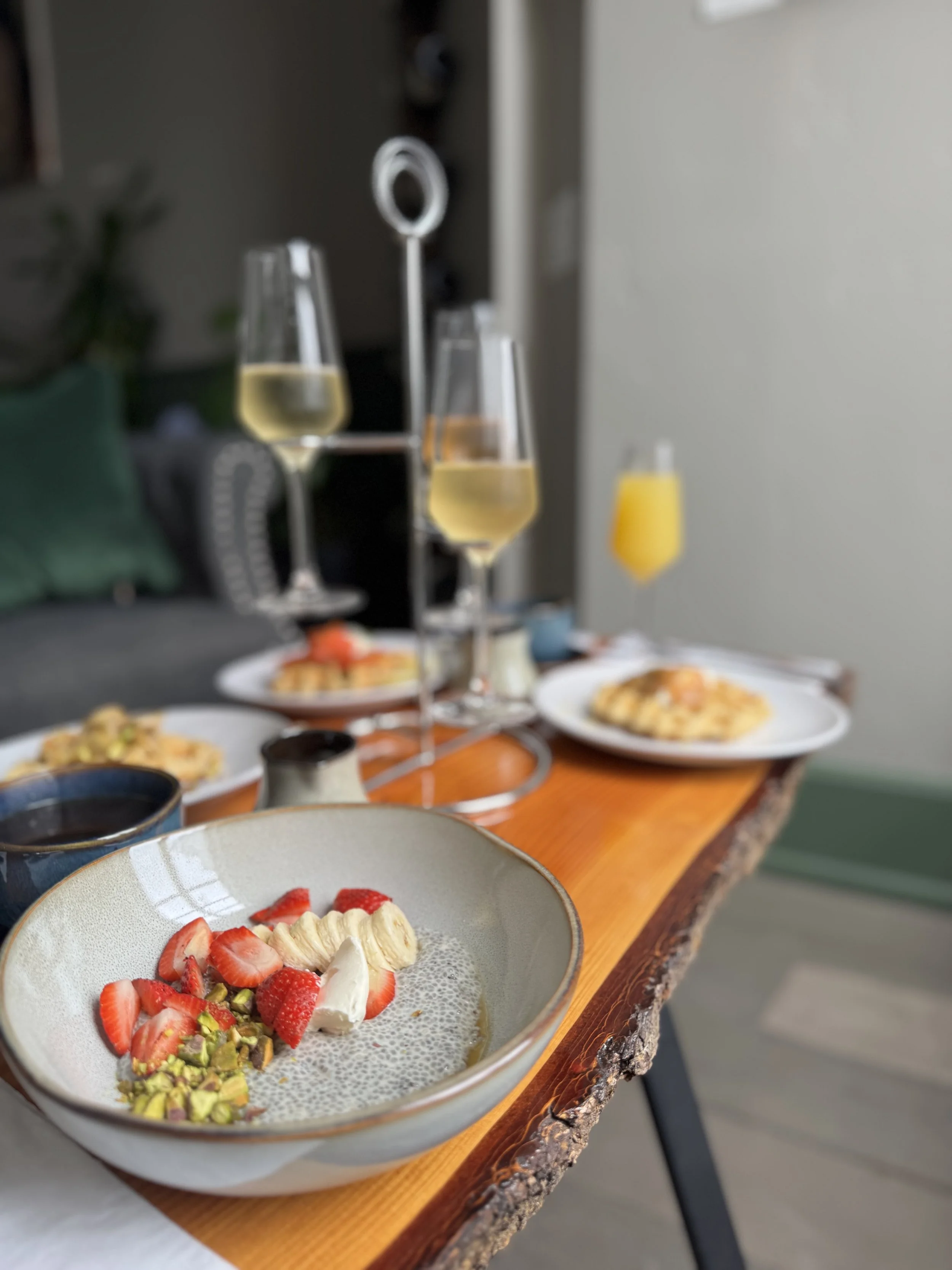 Table set with brunch food. Chia seed pudding with berries, pistachios and cream in a bowl, sparkling wine tower in background, blue coffee cup, and three gluten free waffles in background