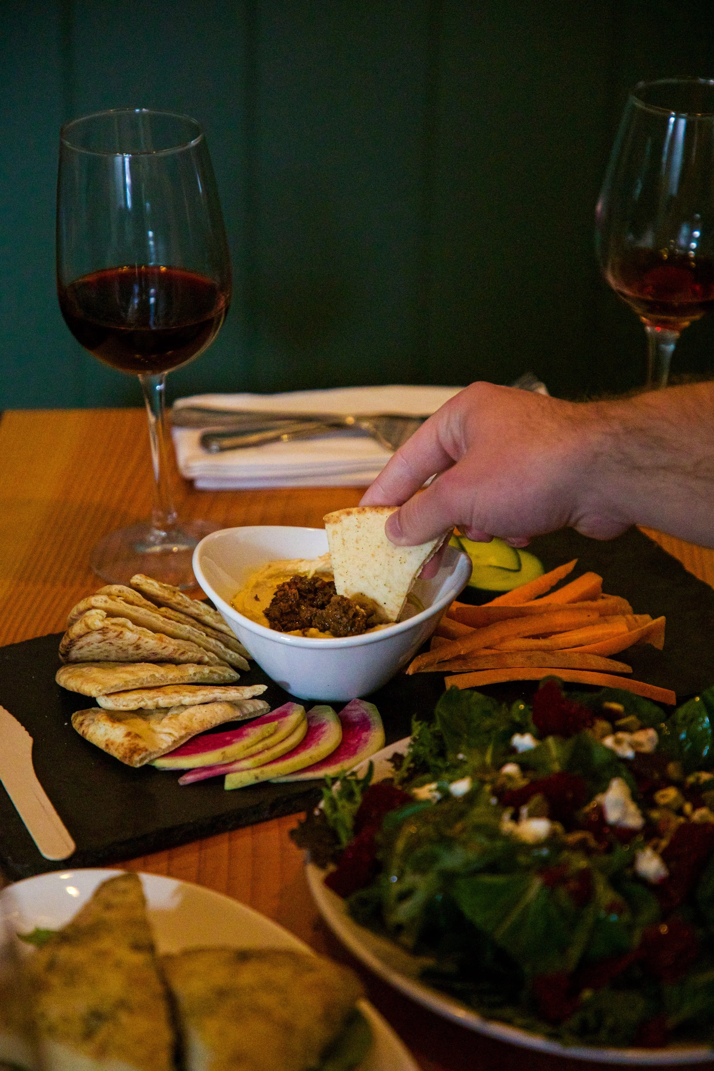 A dining table with two glasses of red wine, a small bowl of hummus, pita bread, sliced radishes, carrots, and a mixed green salad with goat cheese and beets.