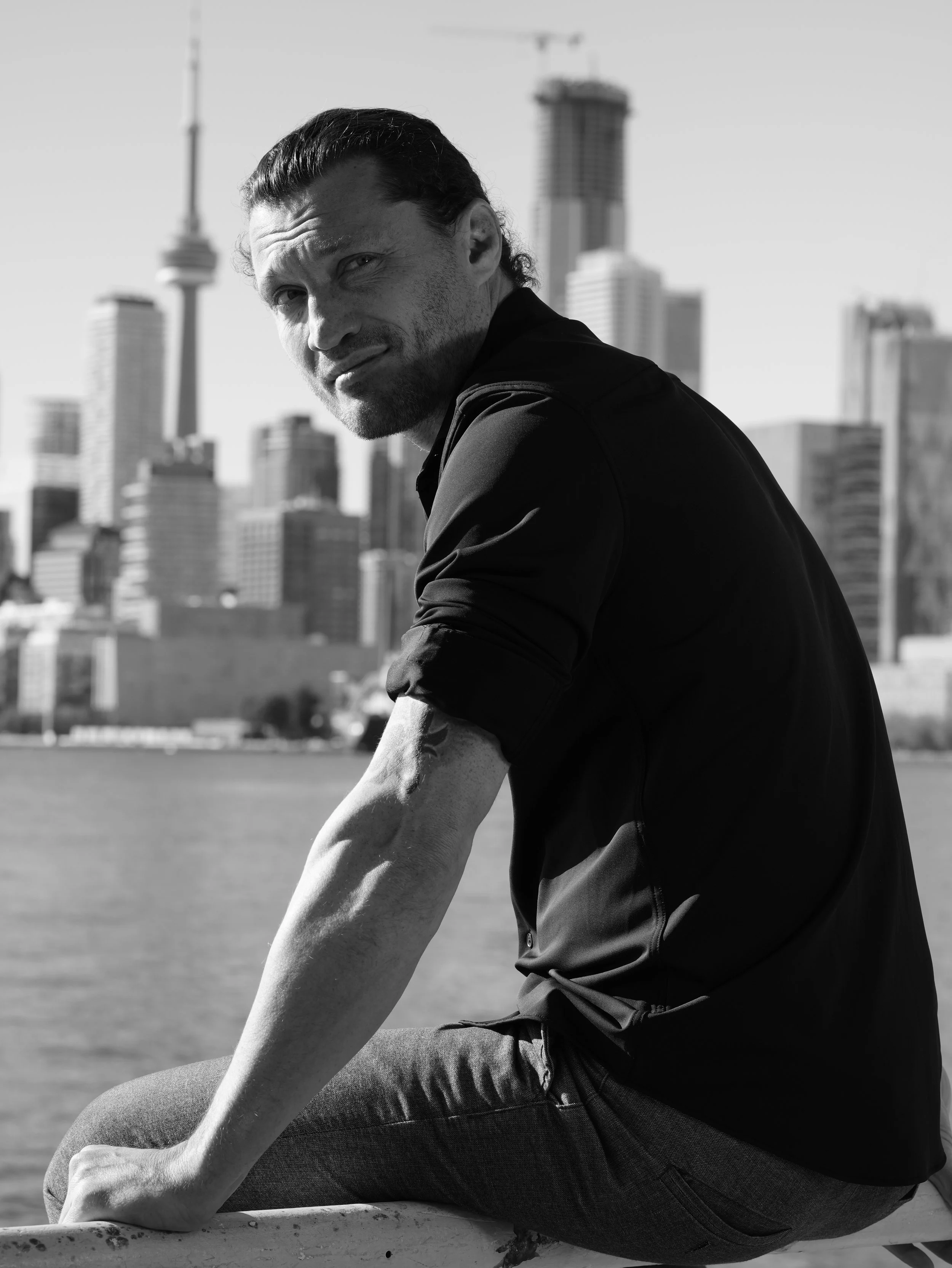 A man with curly hair and a beard standing on a city street at night, illuminated by bright neon signs and billboards.