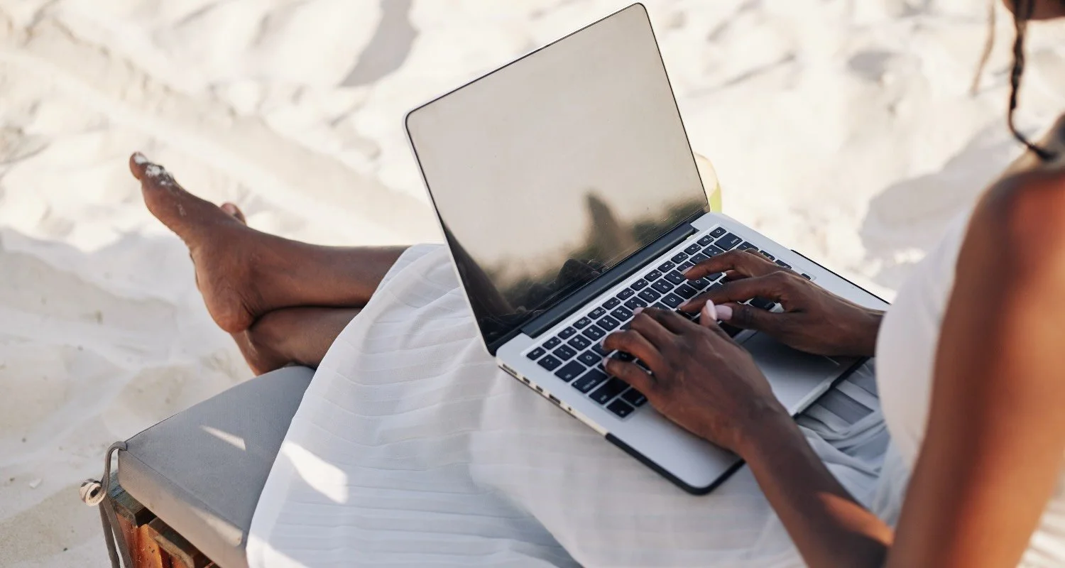 Person sitting on a beach chair using a laptop, with sand visible in the background.