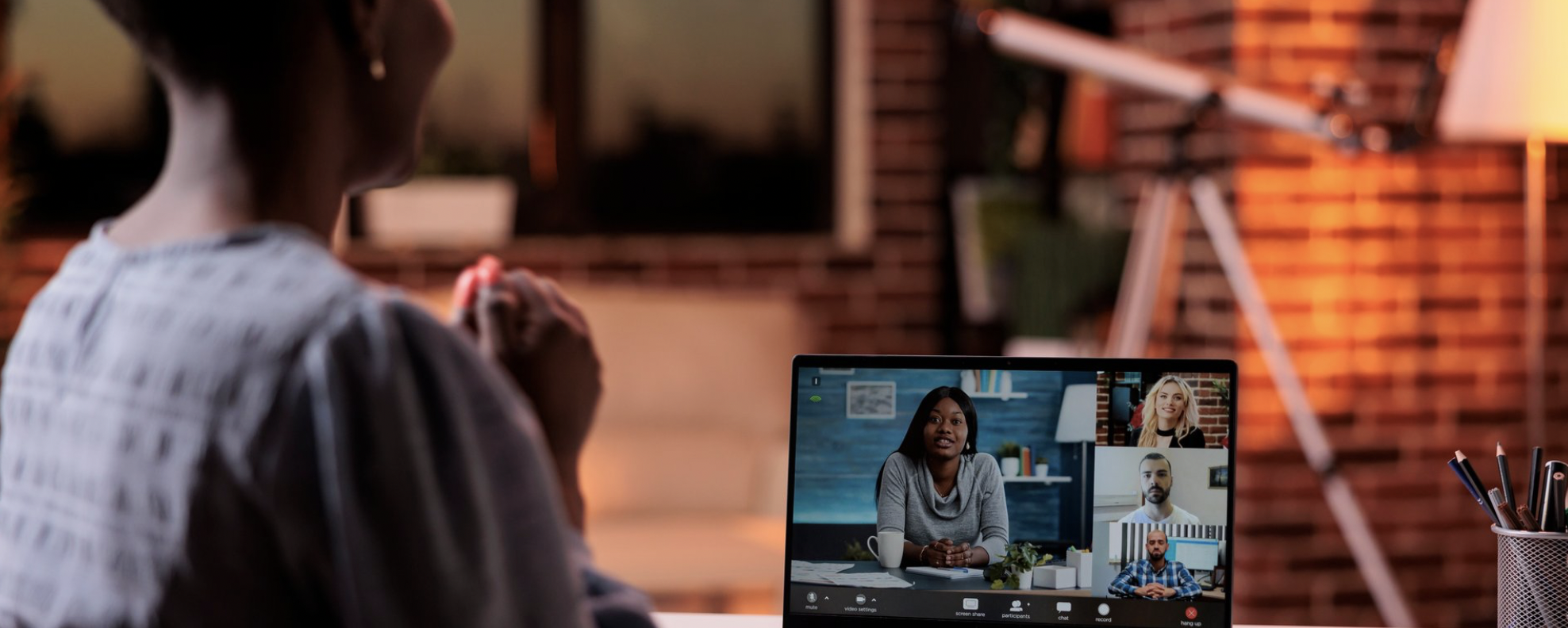 Person participating in a video conference call on a laptop in an indoor setting with warm lighting and a brick wall in the background