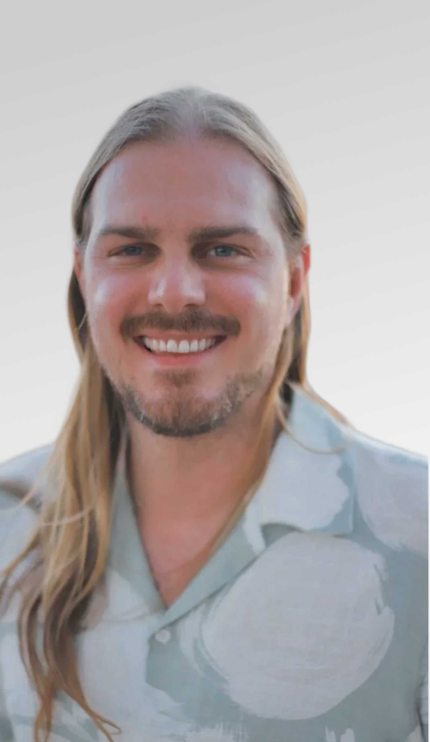 Close-up of a smiling man with long hair, a beard, and a light-colored shirt, against a plain background.
