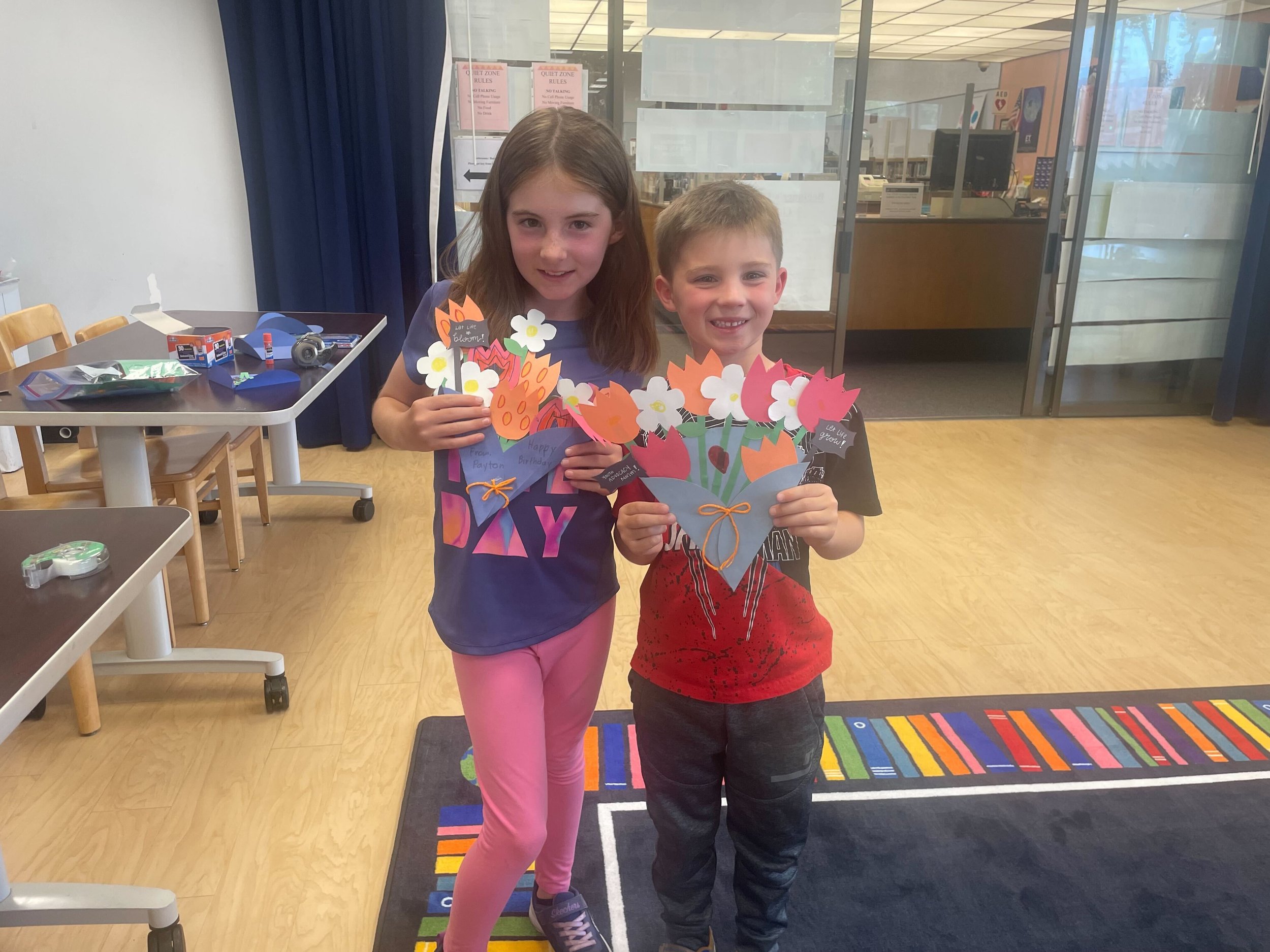 Two children holding paper flower crafts in a classroom setting.