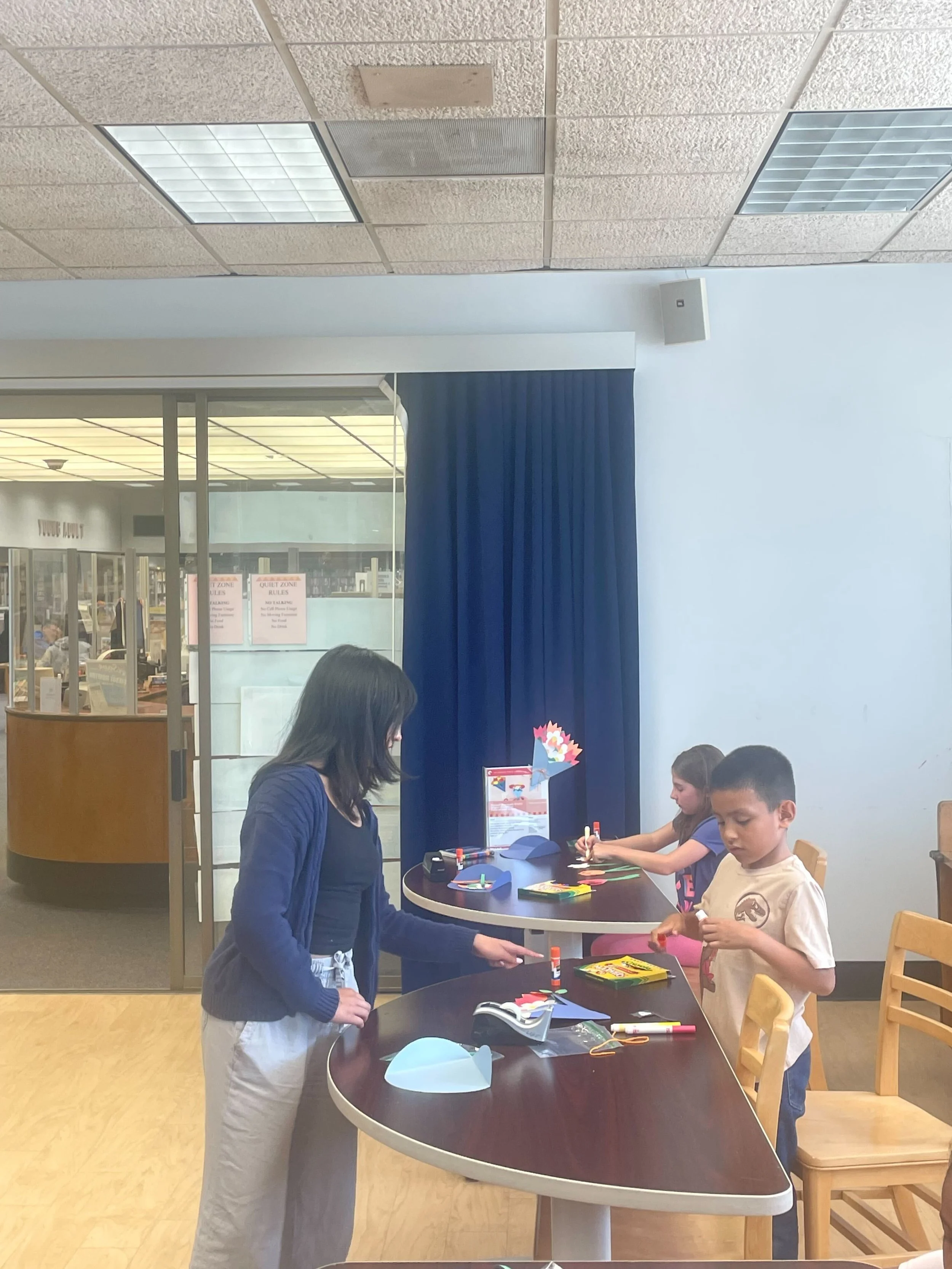 Children and an adult engaged in a craft activity at a library table, with scissors, glue, and paper on the table.
