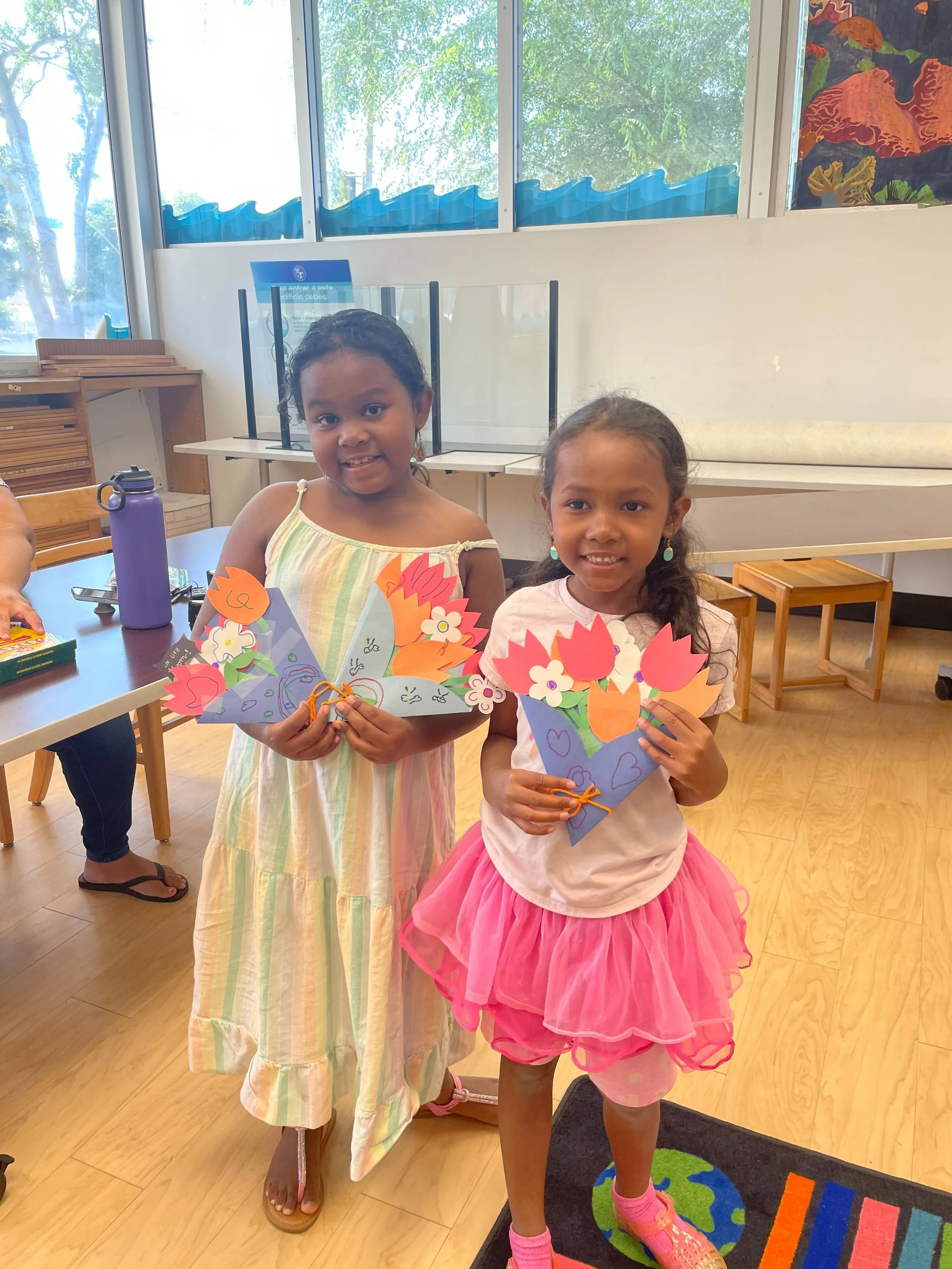 Two young girls in a classroom holding colorful paper crafts shaped like flowers and hearts.