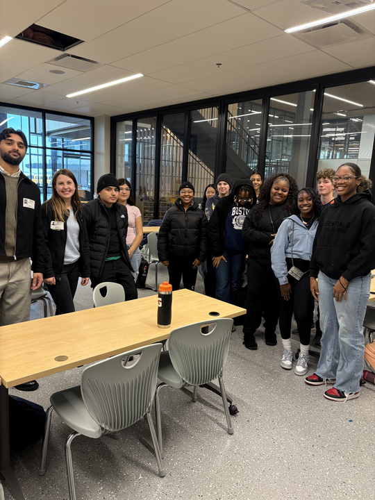Group of young adults standing in a modern office or conference room with glass walls, smiling at the camera.