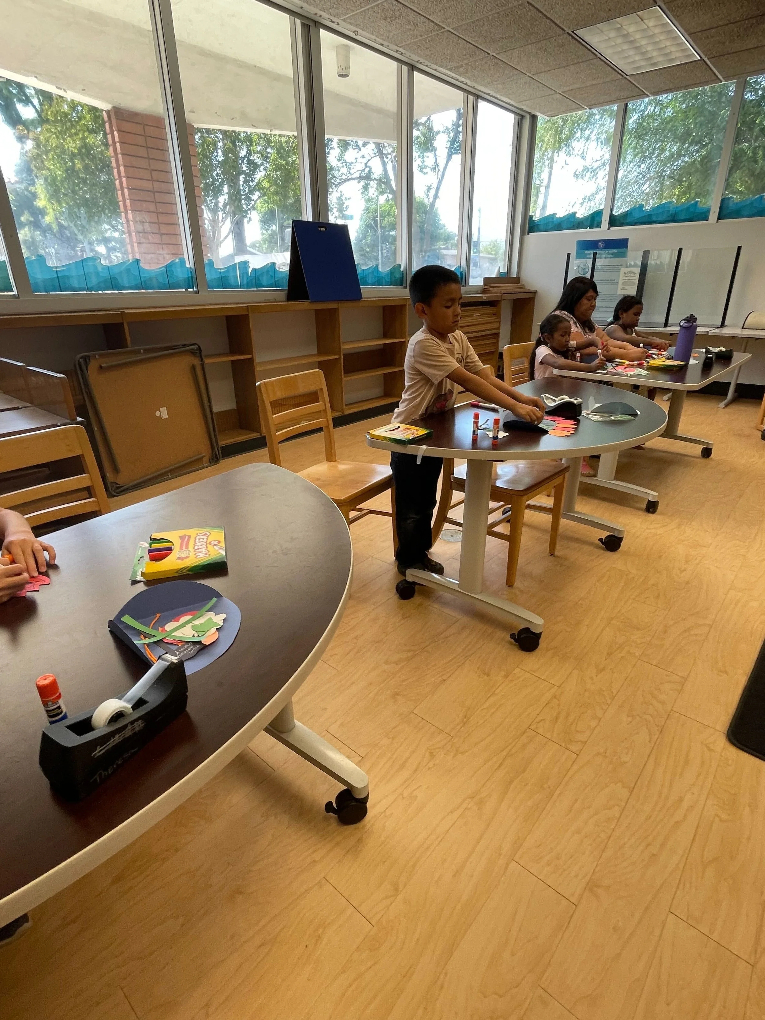 Children doing crafts at tables in a classroom with large windows and natural light.