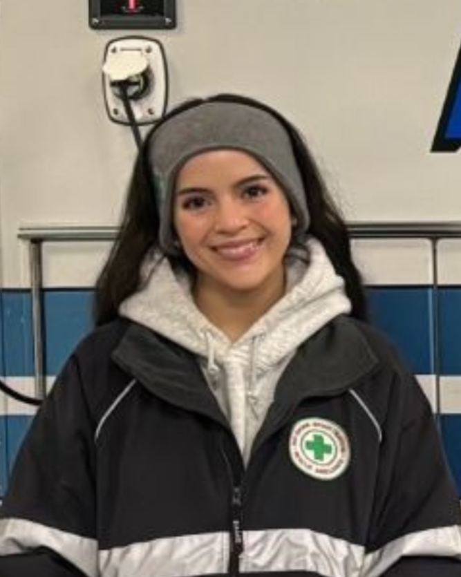A young woman with long dark hair, wearing a gray beanie, white hoodie, and black jacket with an EMT emblem, smiling against an ambulance.