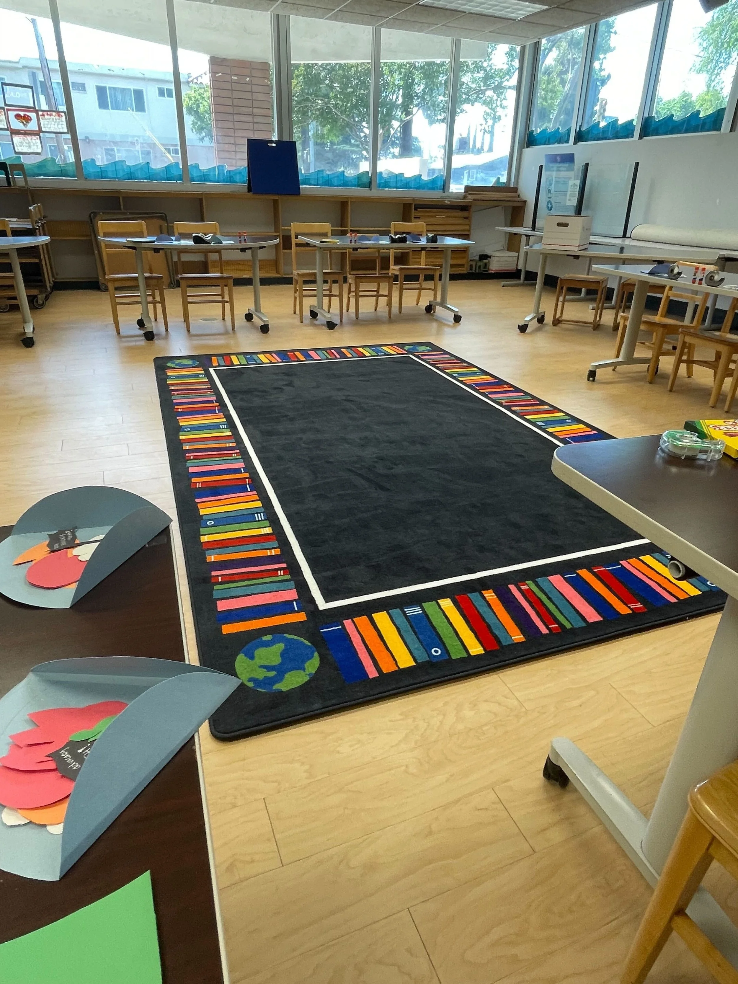 Classroom with tables, chairs, and a colorful bordered rug on the floor, large windows in the background, and paper crafts on a table.