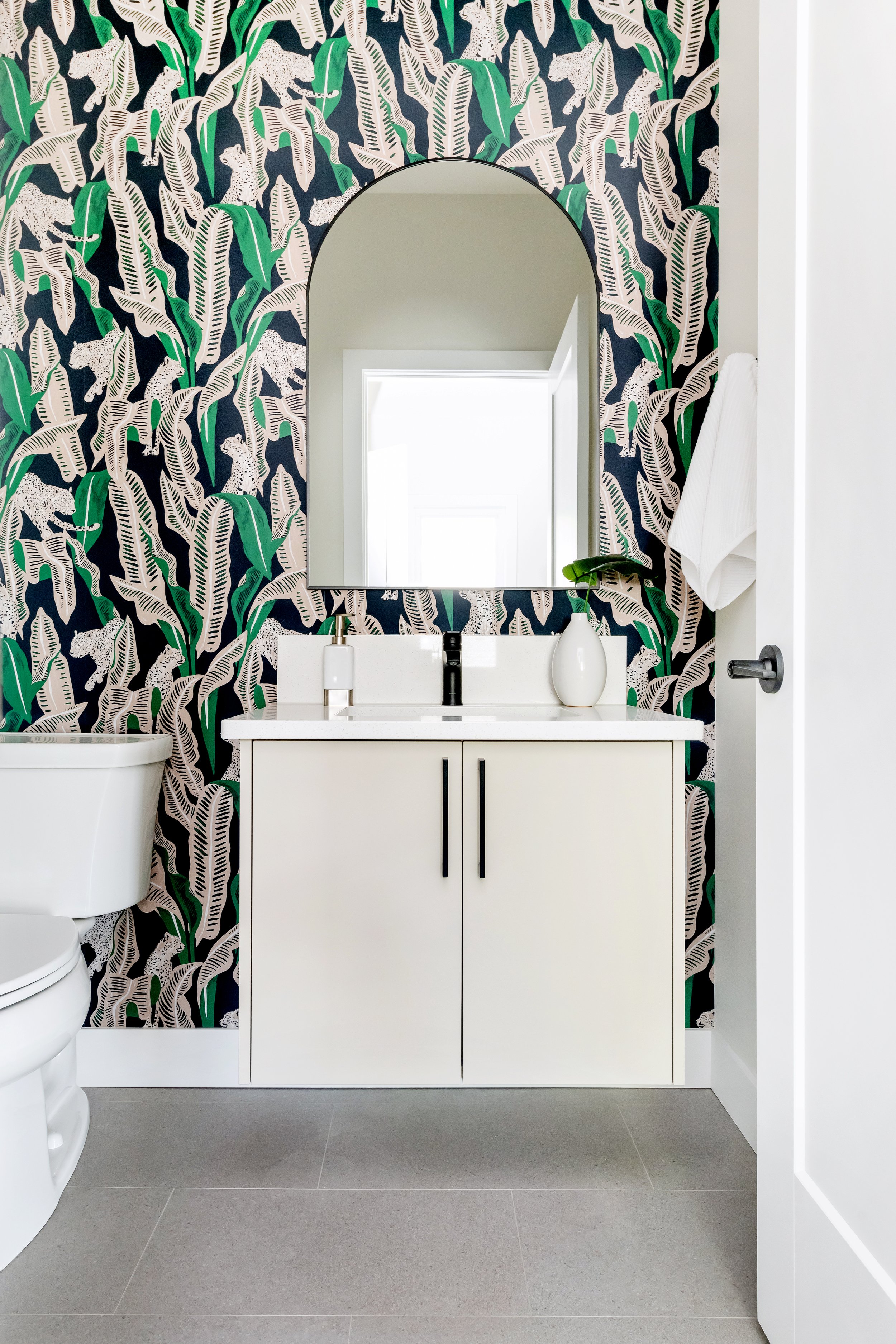 Modern bathroom designed by Bella Vista Developments in Saskatoon, with a vanity, mirror, and a tropical-patterned wallpaper featuring green and white leaves on a dark background, with a white towel hanging on the wall.