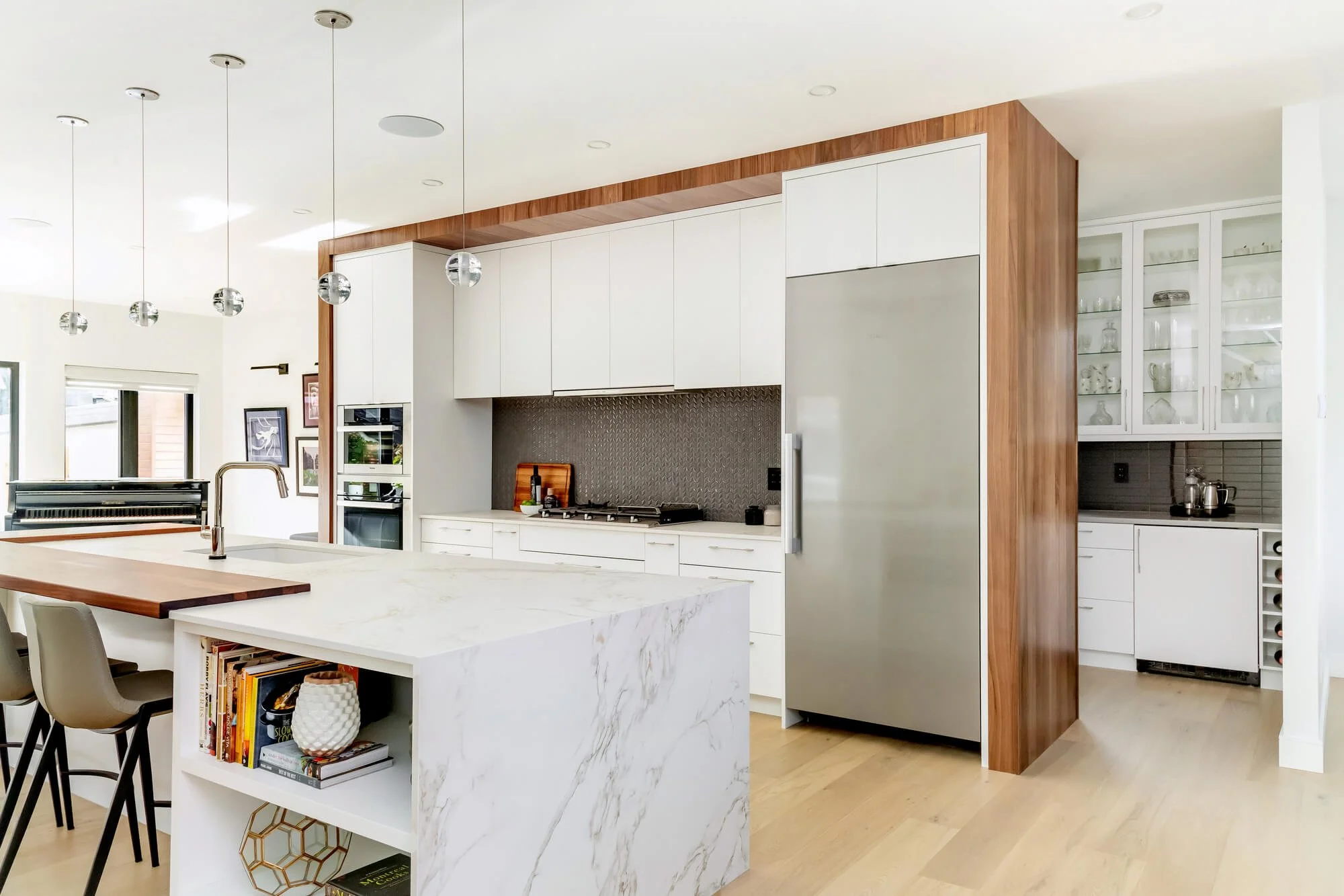 Modern kitchen with white cabinetry, marble island, stainless steel refrigerator, and wood accents, including a curved wood wall and wood flooring.