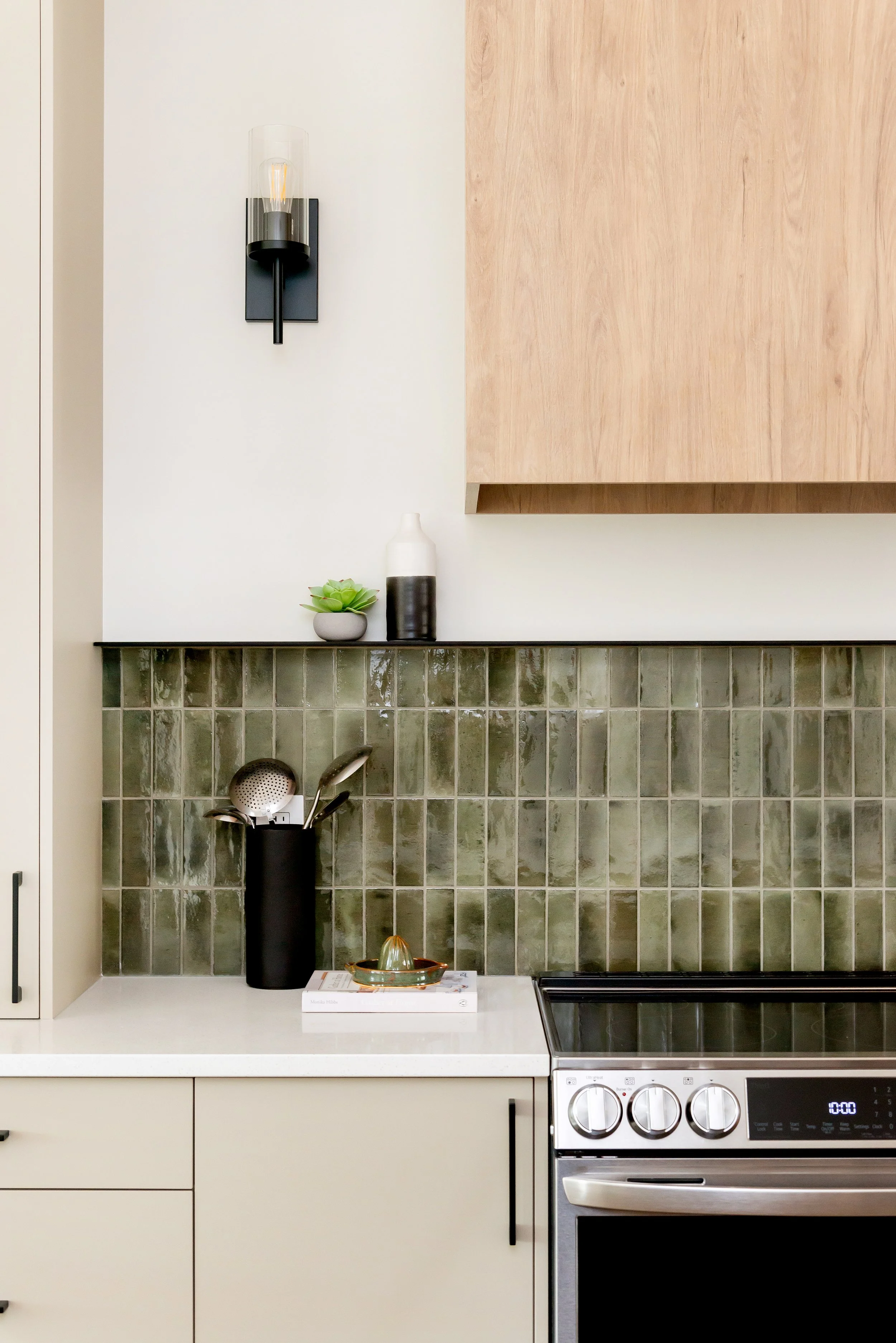 Modern kitchen with beige cabinetry, green tiled backsplash, white countertop, black utensil holder with kitchen tools, decorative vase and small plant on the shelf, wall-mounted light fixture, and stainless steel oven.