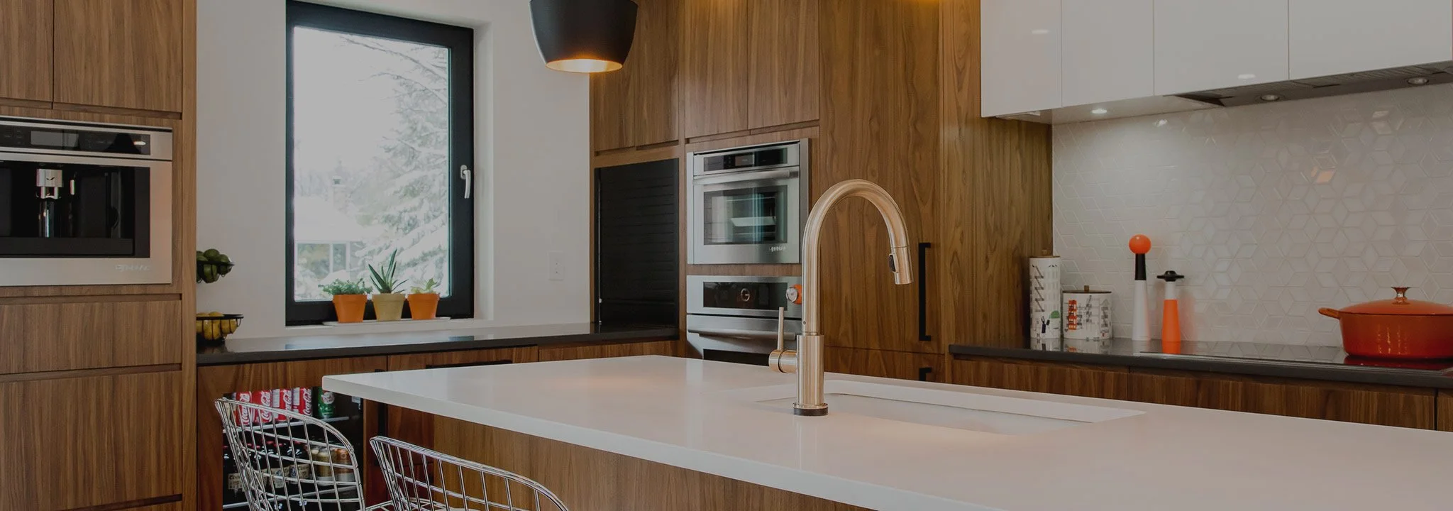 Modern kitchen with wooden cabinets, stainless steel appliances, and a white countertop island with a built-in sink. There are small potted plants on the window sill and various kitchen utensils and decorative items on the counter.