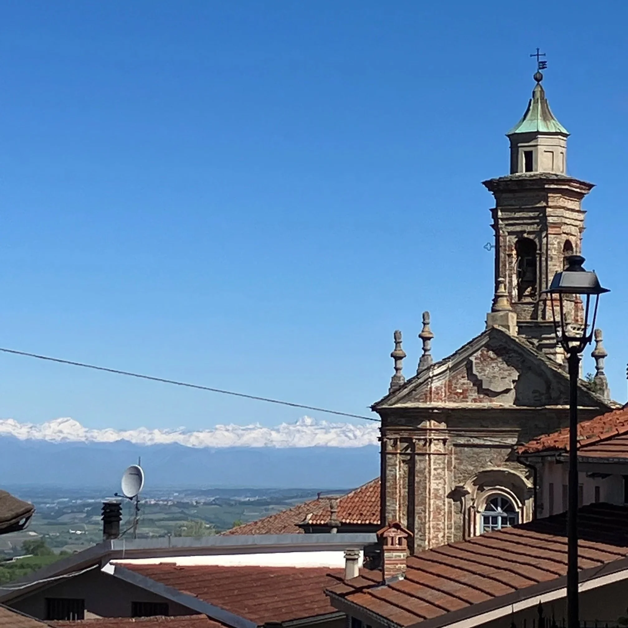 Een van de kerken van Murazzano (Alta Langa, Piemonte) met een toren en een klok, omgeven door huizen met rode dakpannen, met een landschap en bergen op de achtergrond onder een blauwe hemel.