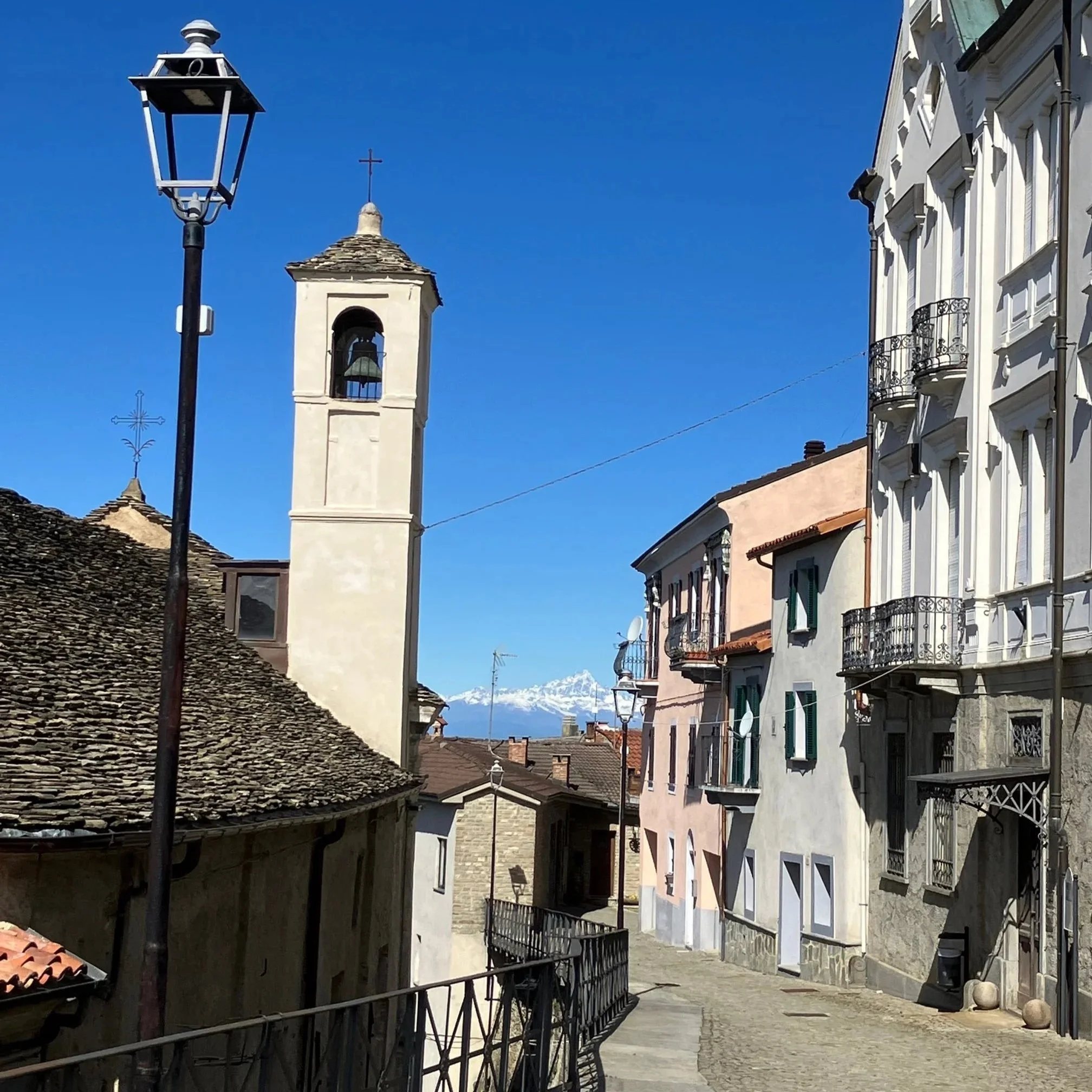 Smal straatje in Murazzano (Alta Langa, Piemonte) met kleurrijke gebouwen en een kerktoren, met bergen op de achtergrond onder een heldere blauwe hemel.