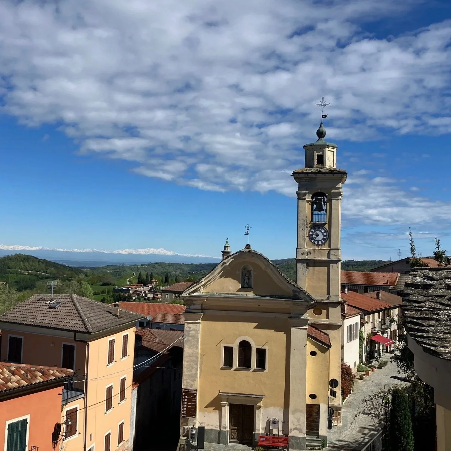 Uitzicht op een plein in Murazzano (Alta Langa, Piemonte) met kerktoren en huizen in een bergachtig landschap onder een blauwe hemel met wolken.