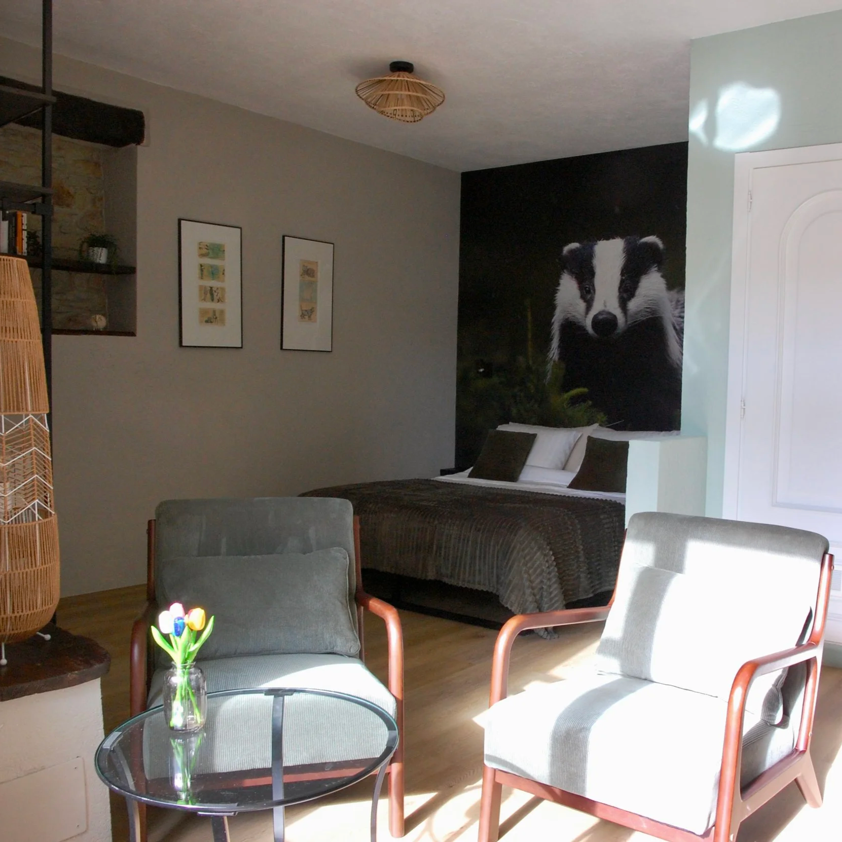 Interieur van een slaapkamer met een bed, groene kussens, een muurposter van een das, zwarte fauteuils en een glazen tafel, met decoratieve elementen en een hanglamp.